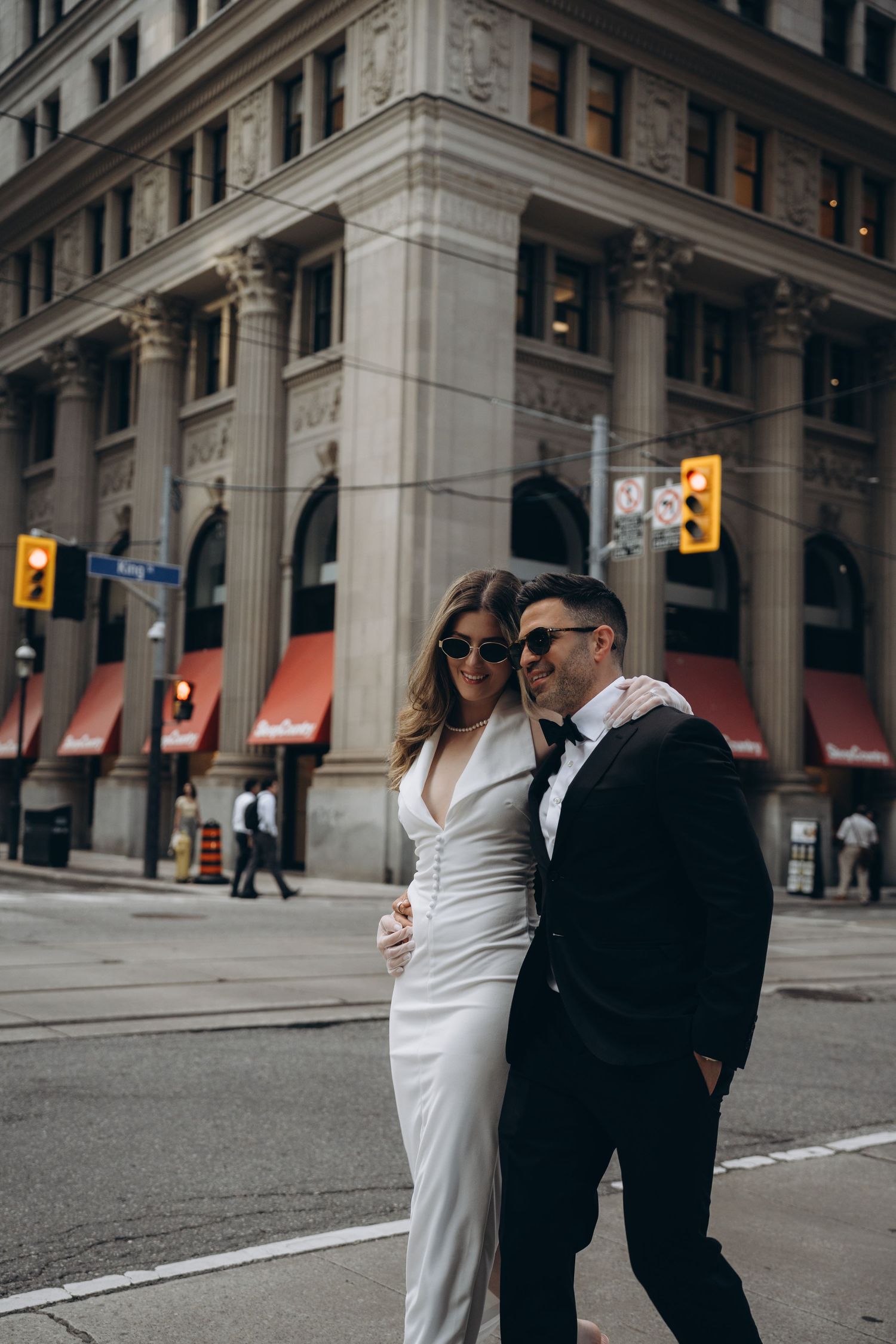 A couple in formal attire walks along a city street with classic architecture and traffic lights in the background.