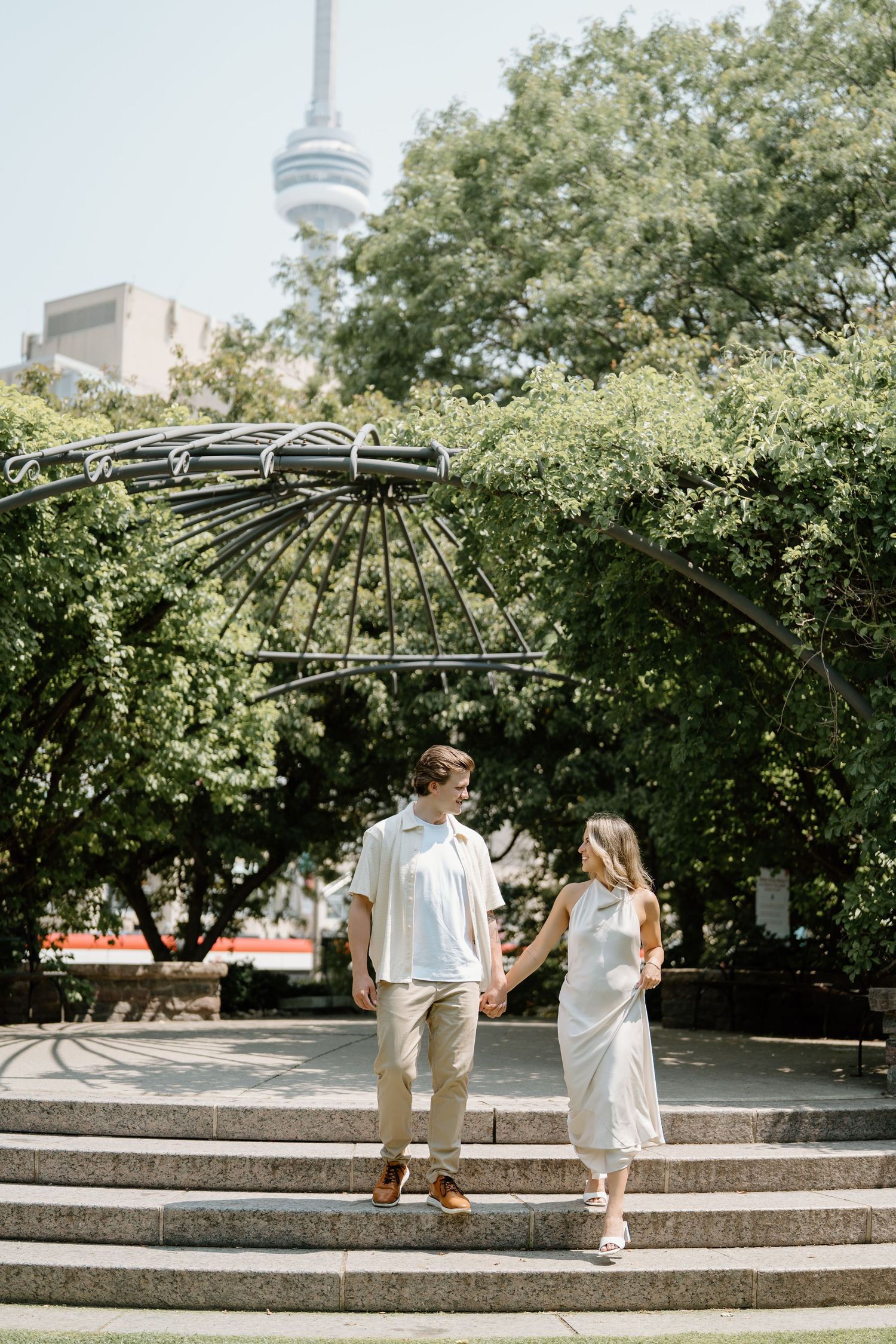 A couple poses on garden steps under a decorative metal archway with the CN Tower rising behind them in Toronto.