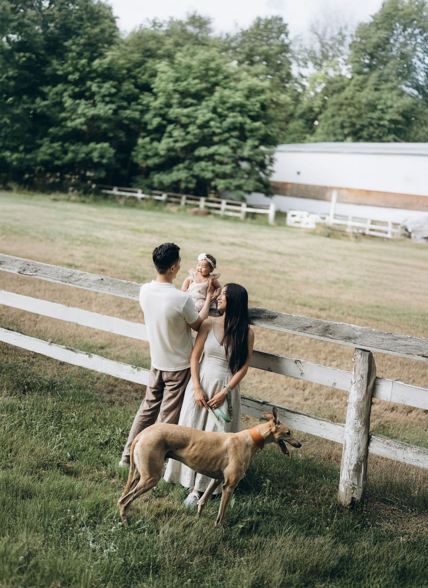 A family enjoying a summer day by a white wooden fence on a farm with their brown dog.