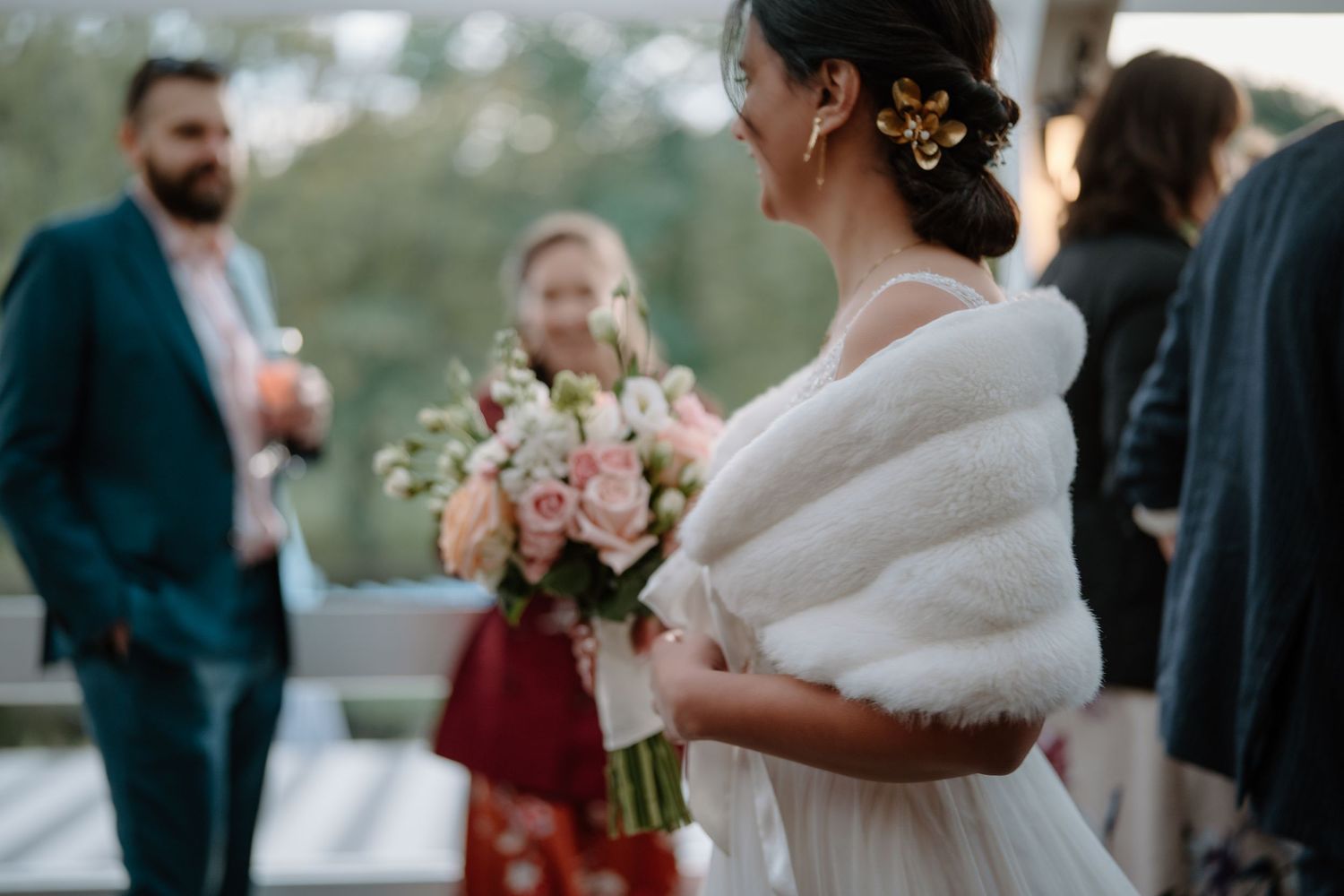 A bride in a white dress and fur wrap holds a pink bouquet during an outdoor reception.