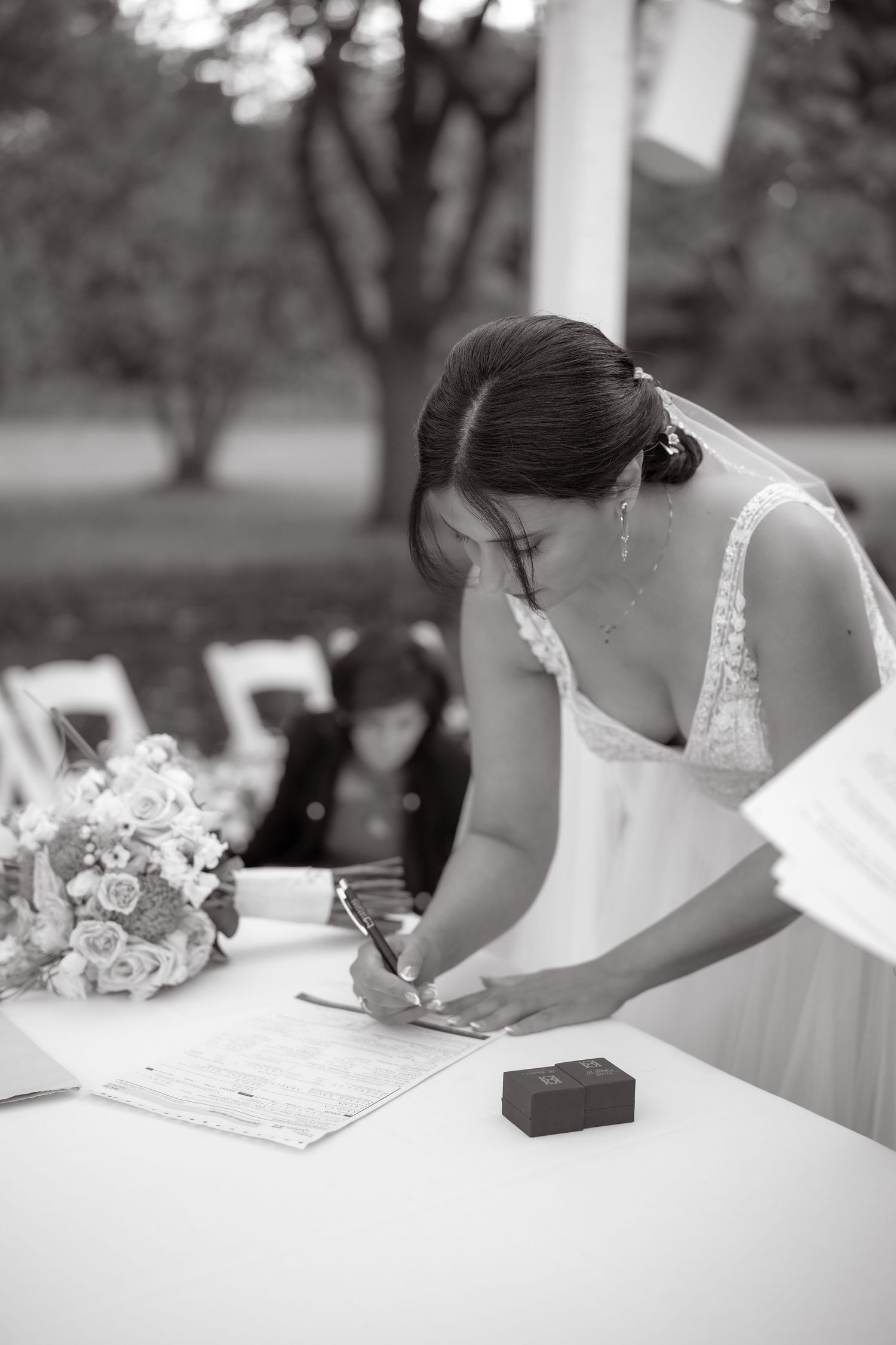 Bride in white dress signing wedding documents at outdoor ceremony with bouquet on table.