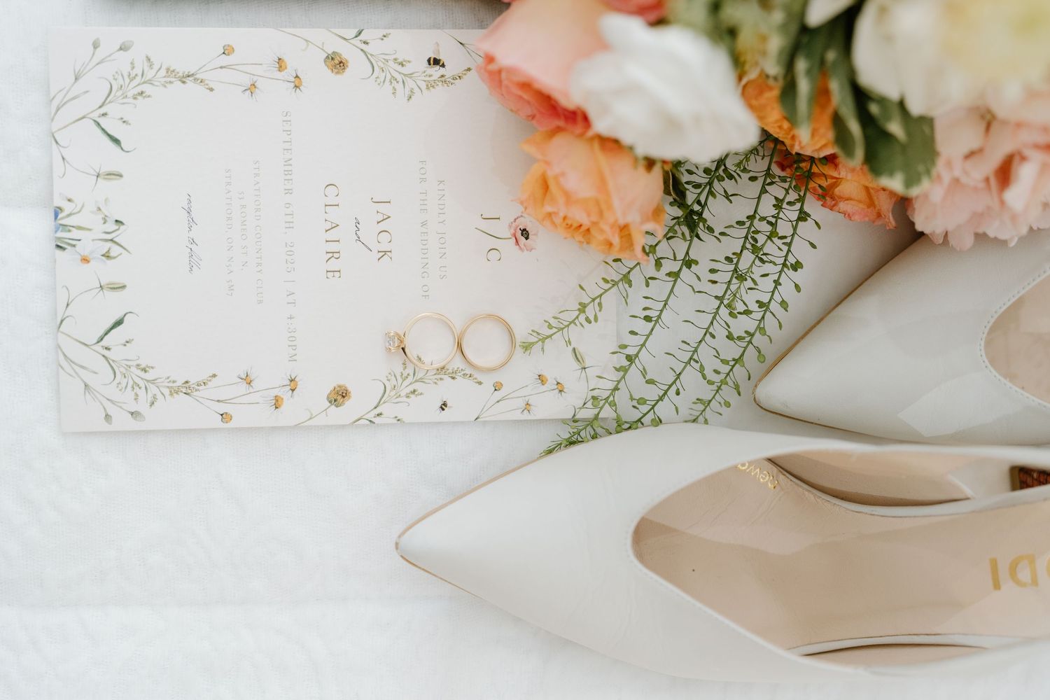Wedding details including elegant white heels, delicate flowers, and a decorative invitation card on a light background.