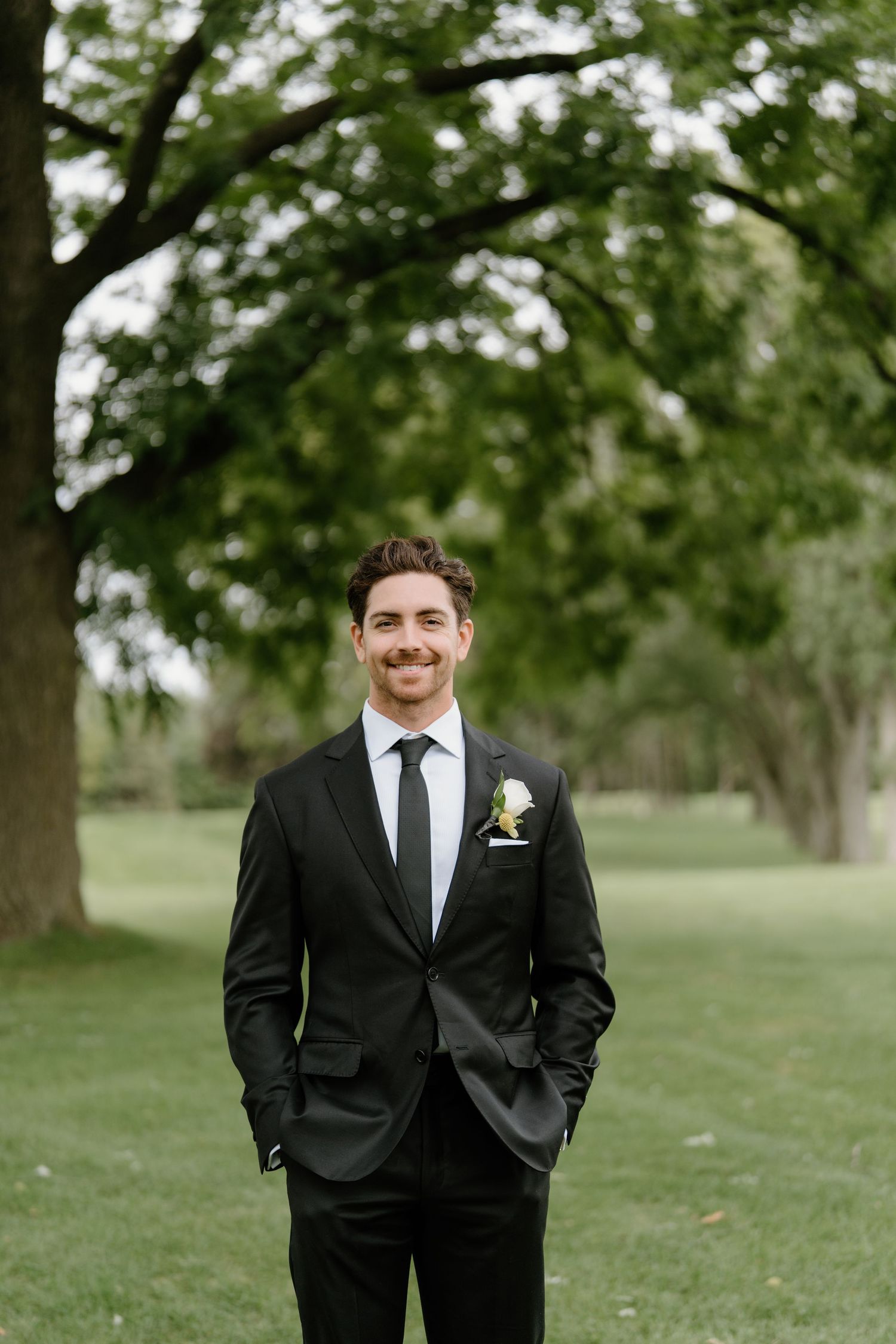 Groom in classic black tuxedo poses confidently on green lawn.
