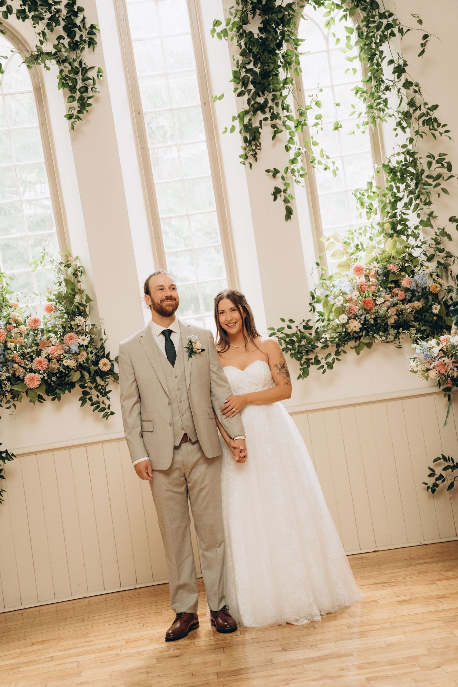 Wedding couple shares a playful first look moment in a white indoor venue decorated with greenery and pink flowers.