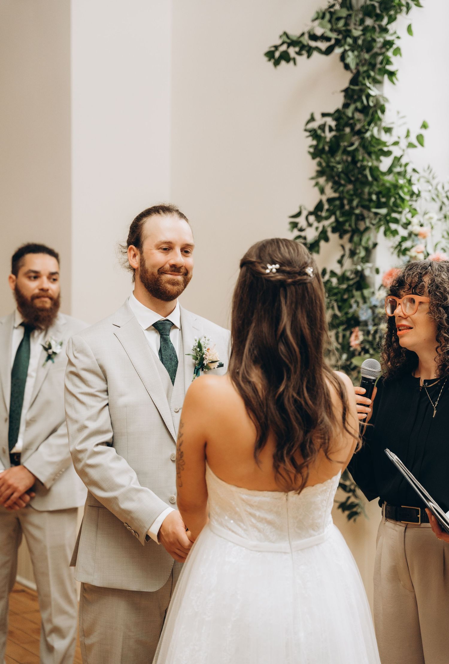 A wedding party stands against a white wall with greenery, sharing joyful moments during a candid photo session.