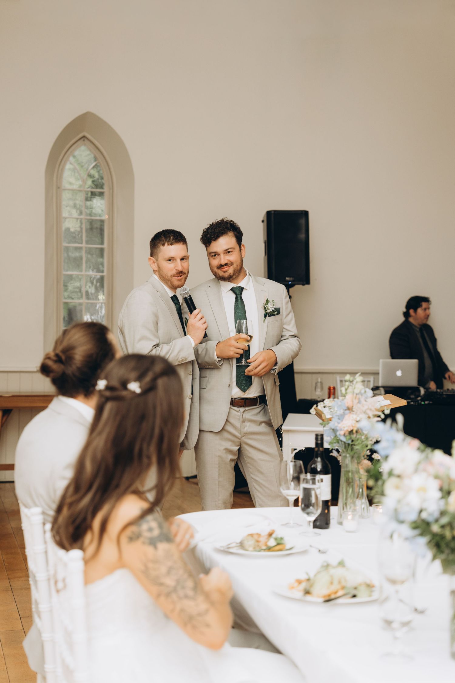 Wedding reception guests seated at elegantly decorated tables with white linens and floral centerpieces in a bright venue.