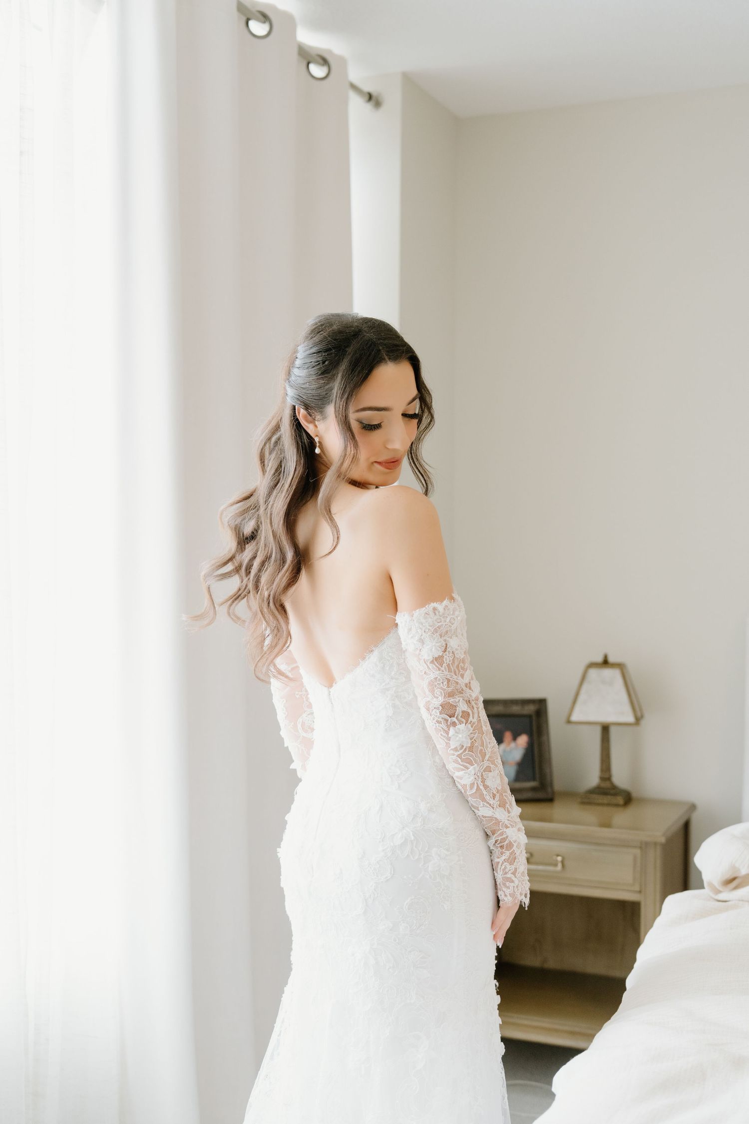 A bride in a white lace wedding gown with long sleeves stands elegantly in a bright, minimalist room.