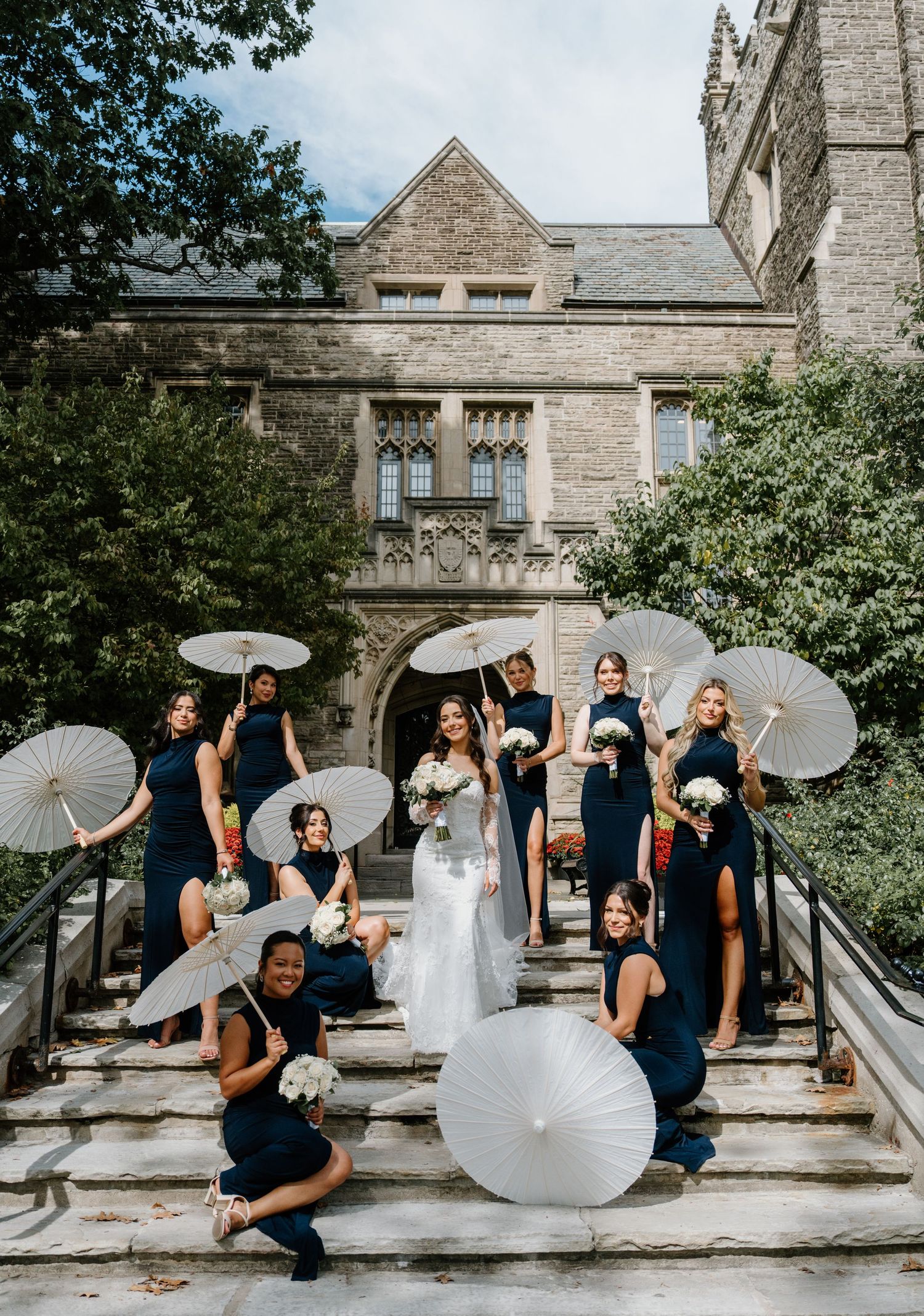 A wedding party in navy blue dresses and white gowns poses playfully with parasols on stone steps of a historic building.