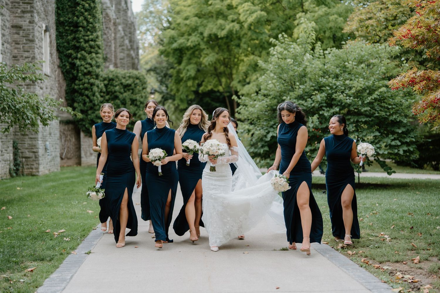 Wedding party in navy blue attire walks together on a garden path surrounded by lush greenery.