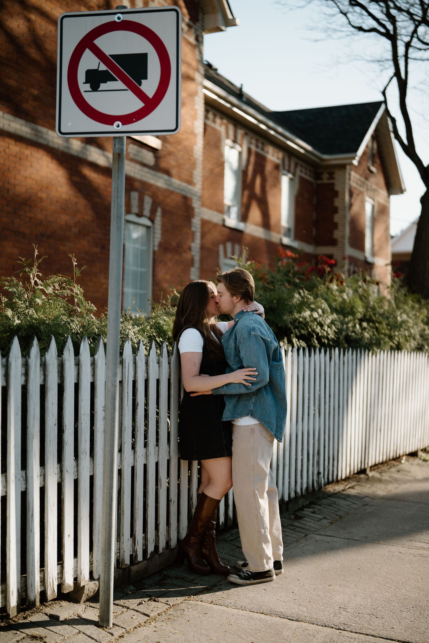 A romantic couple embraces near a white picket fence and street sign on a sunny afternoon.