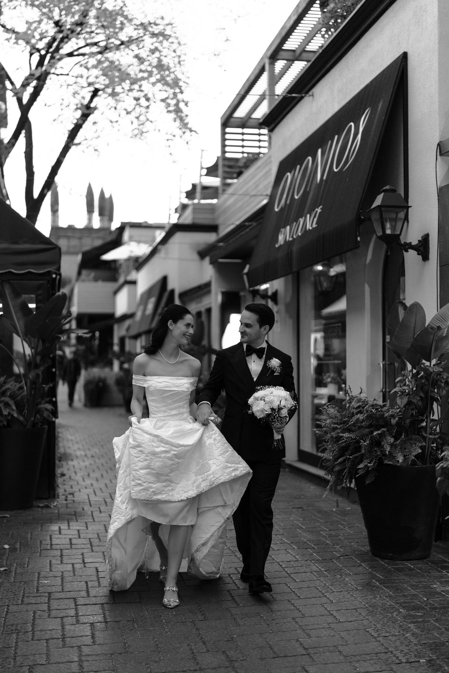 A bride and groom walk along a brick sidewalk outside storefronts in a black and white photo series.