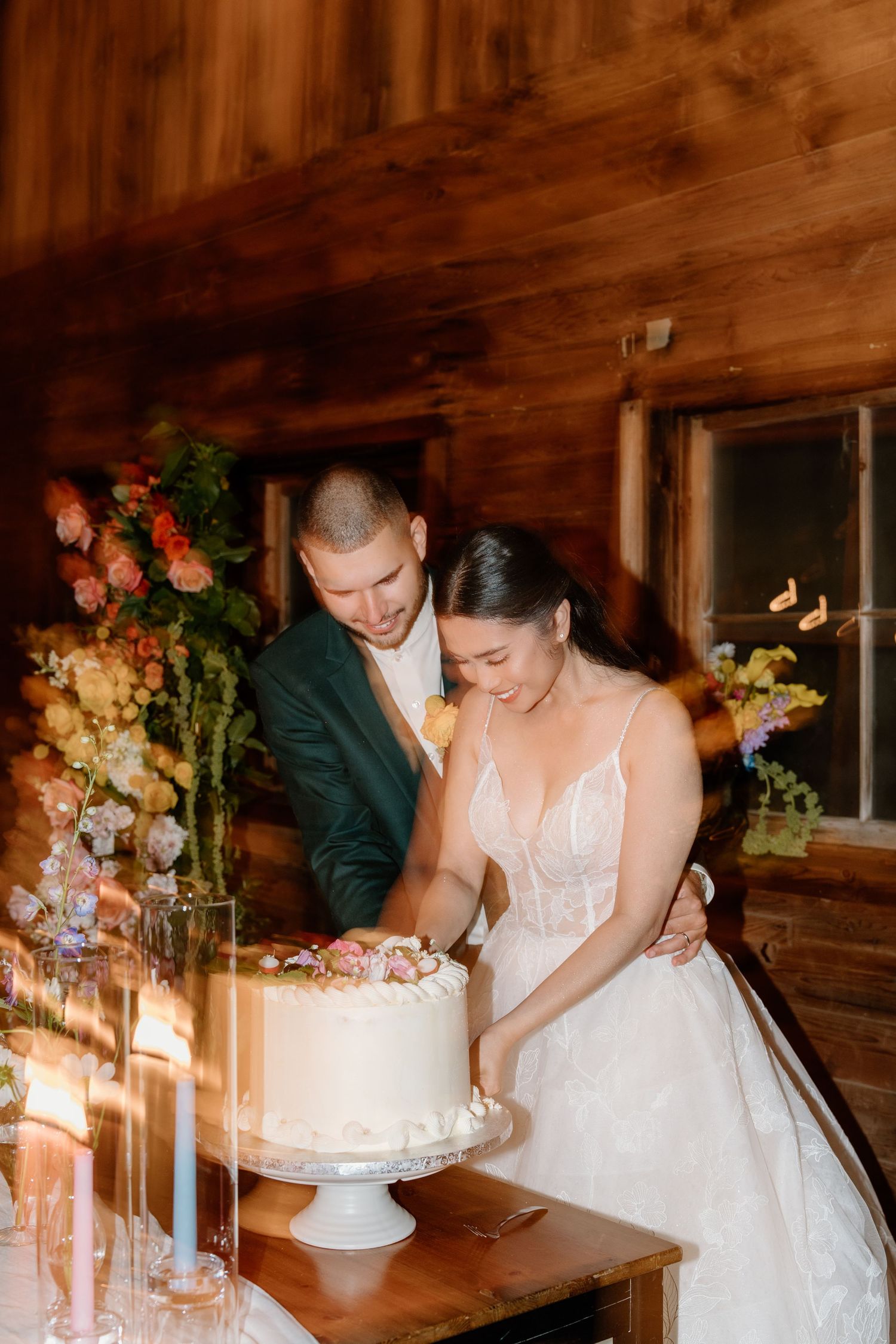 Couple cutting their elegant white wedding cake together in a rustic wooden venue with floral decorations.