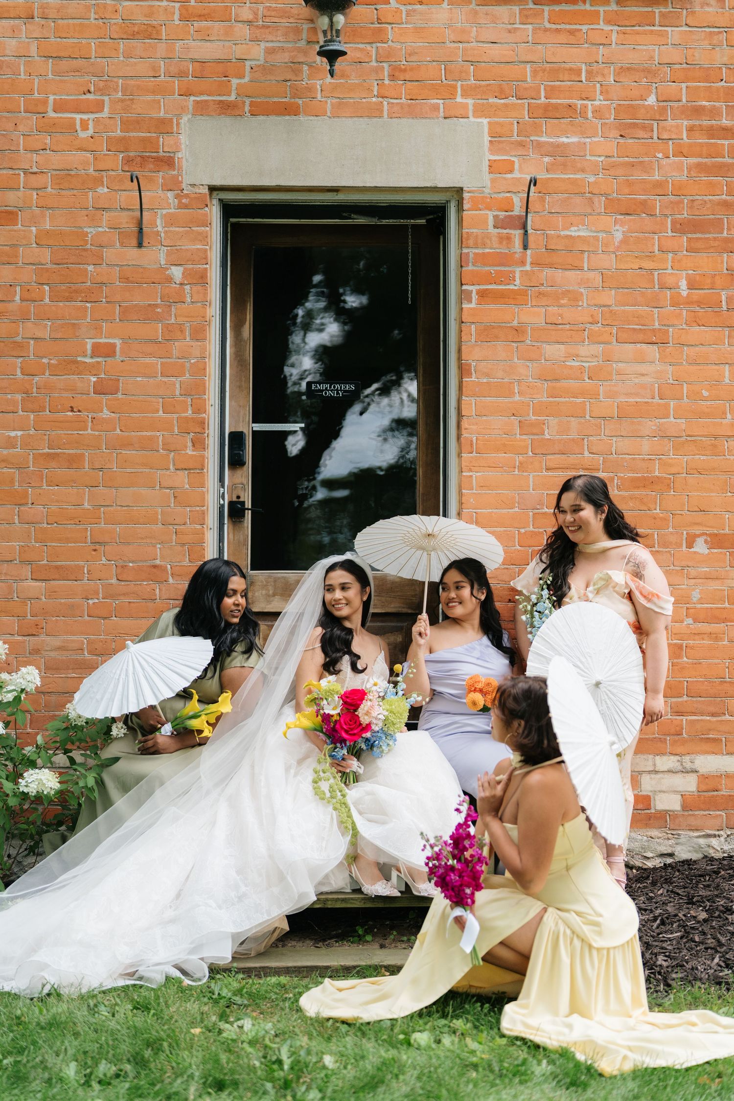 A wedding party in white dresses and yellow bridesmaid dresses poses playfully with flowing veils against a brick building.