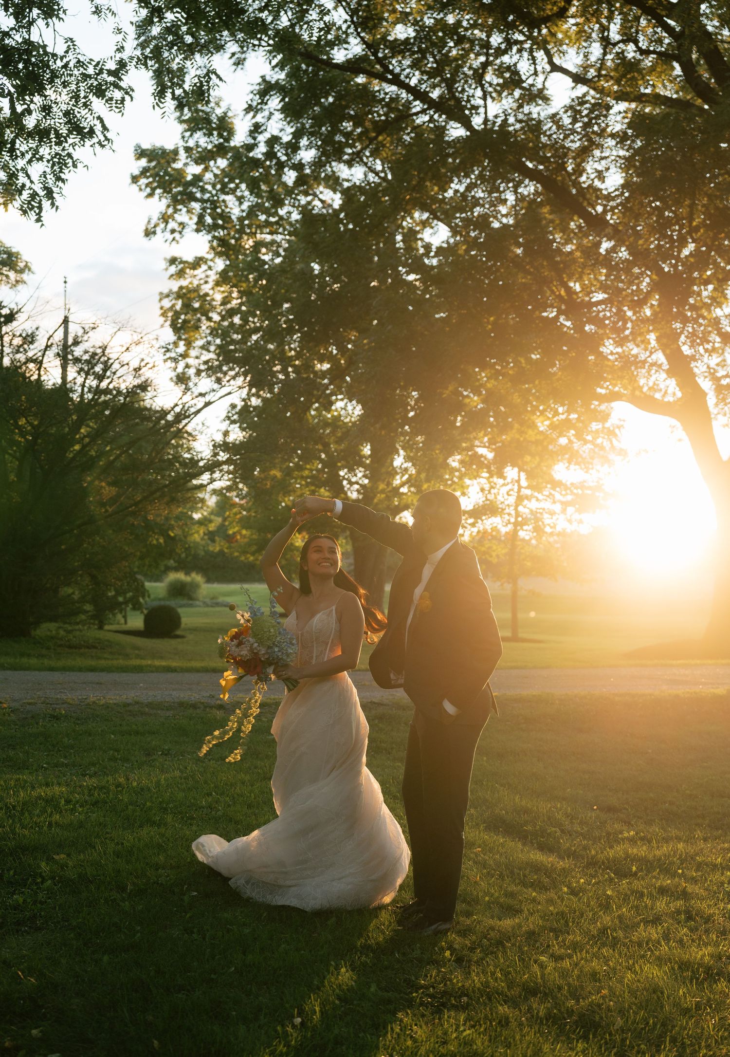 A couple walks through a sunlit park at golden hour as her white dress flows in the breeze.