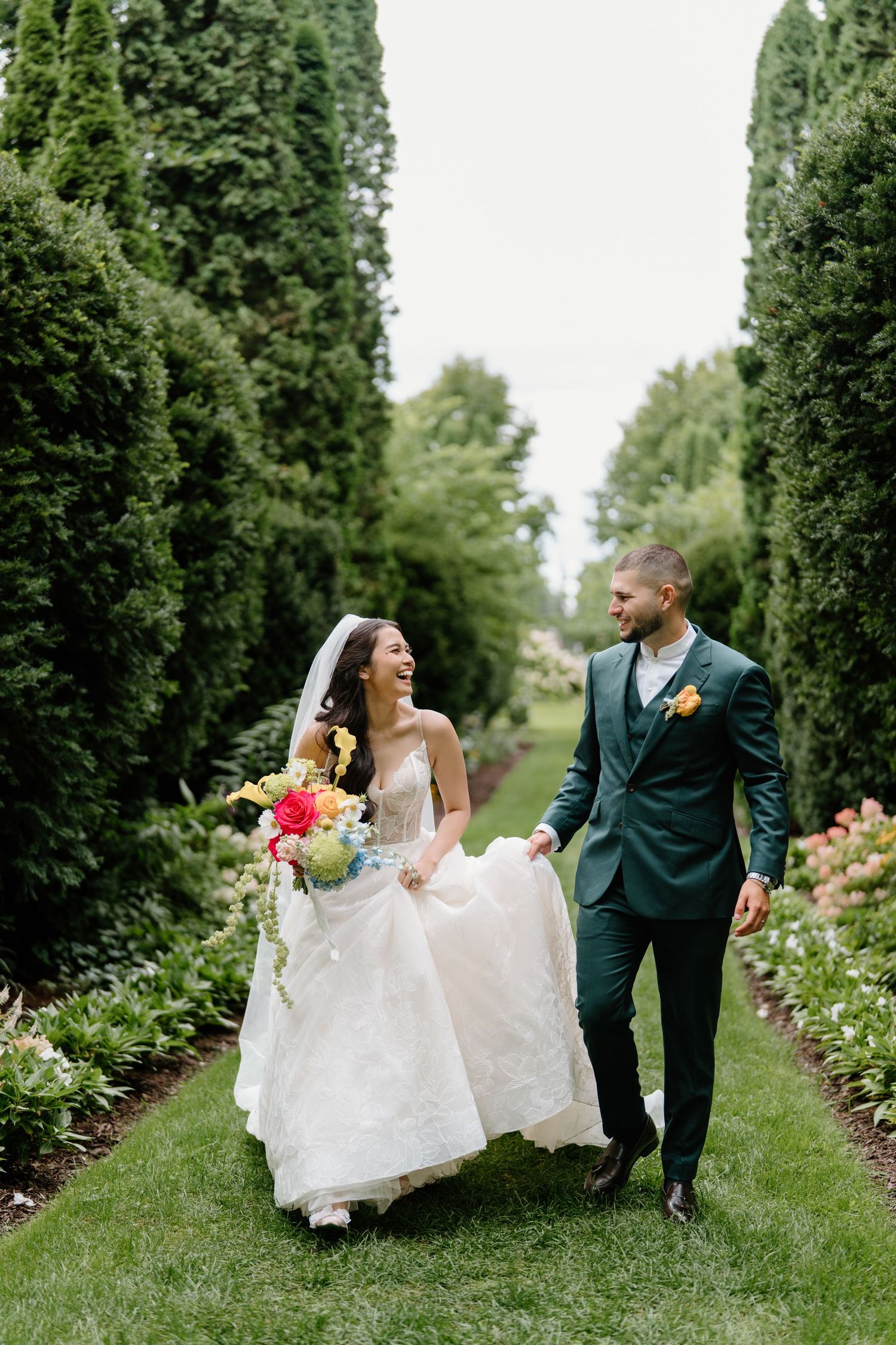 Bride and groom share joyful moment while walking through garden path lined with cypress trees.