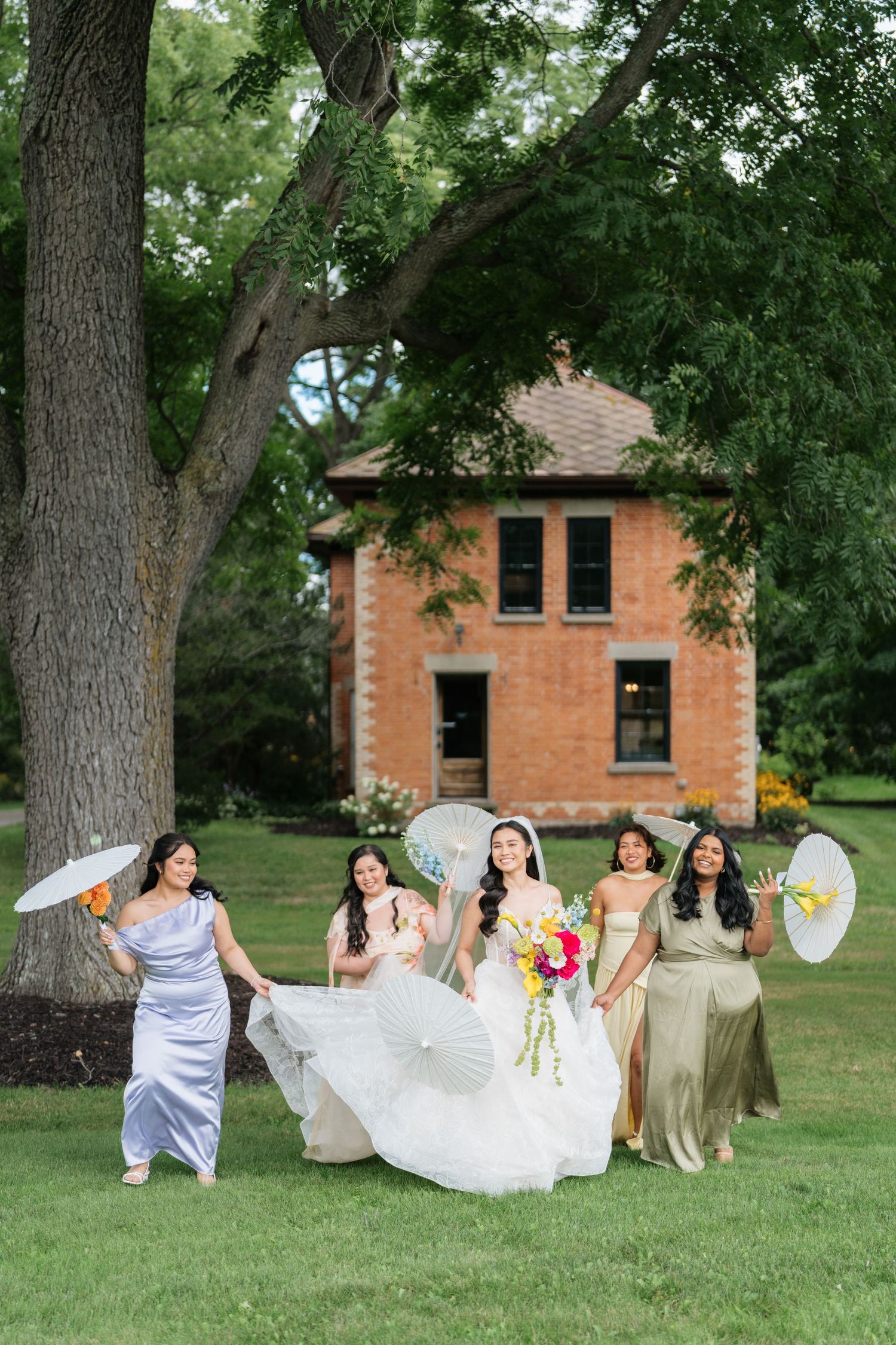 A bridal party poses with decorative parasols on a lawn in front of a rustic brick house surrounded by trees.