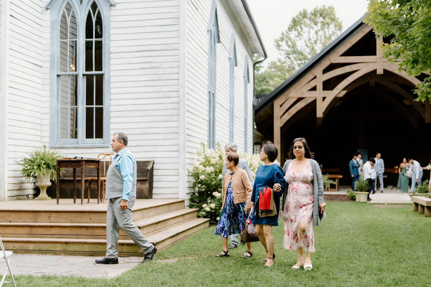 People exit a white chapel with gothic windows and covered entrance on a sunny summer day.