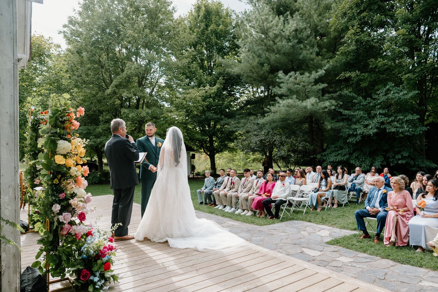 Wedding ceremony takes place outdoors under a covered pavilion with guests seated in white chairs.