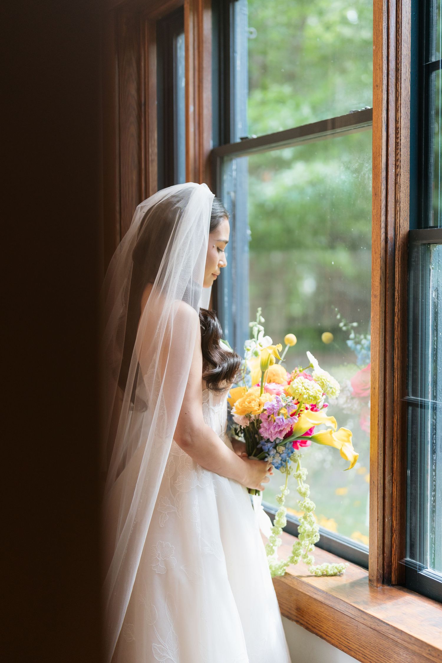 A bride in a white wedding gown and veil gazes thoughtfully out of a sunlit window with colorful flowers in hand.