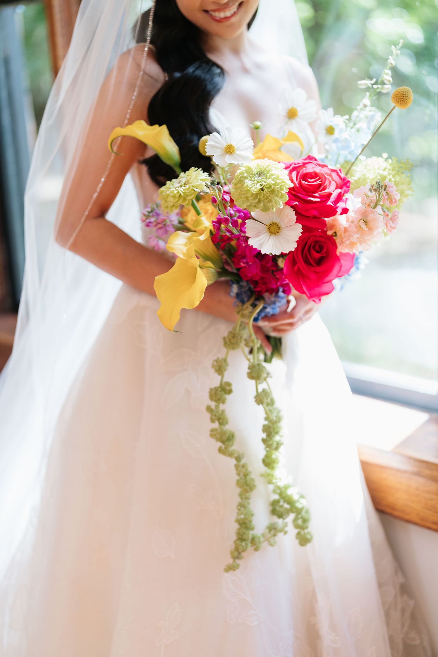 A colorful bridal bouquet features vibrant pink roses, yellow orchids and cascading greenery against a white wedding dress.