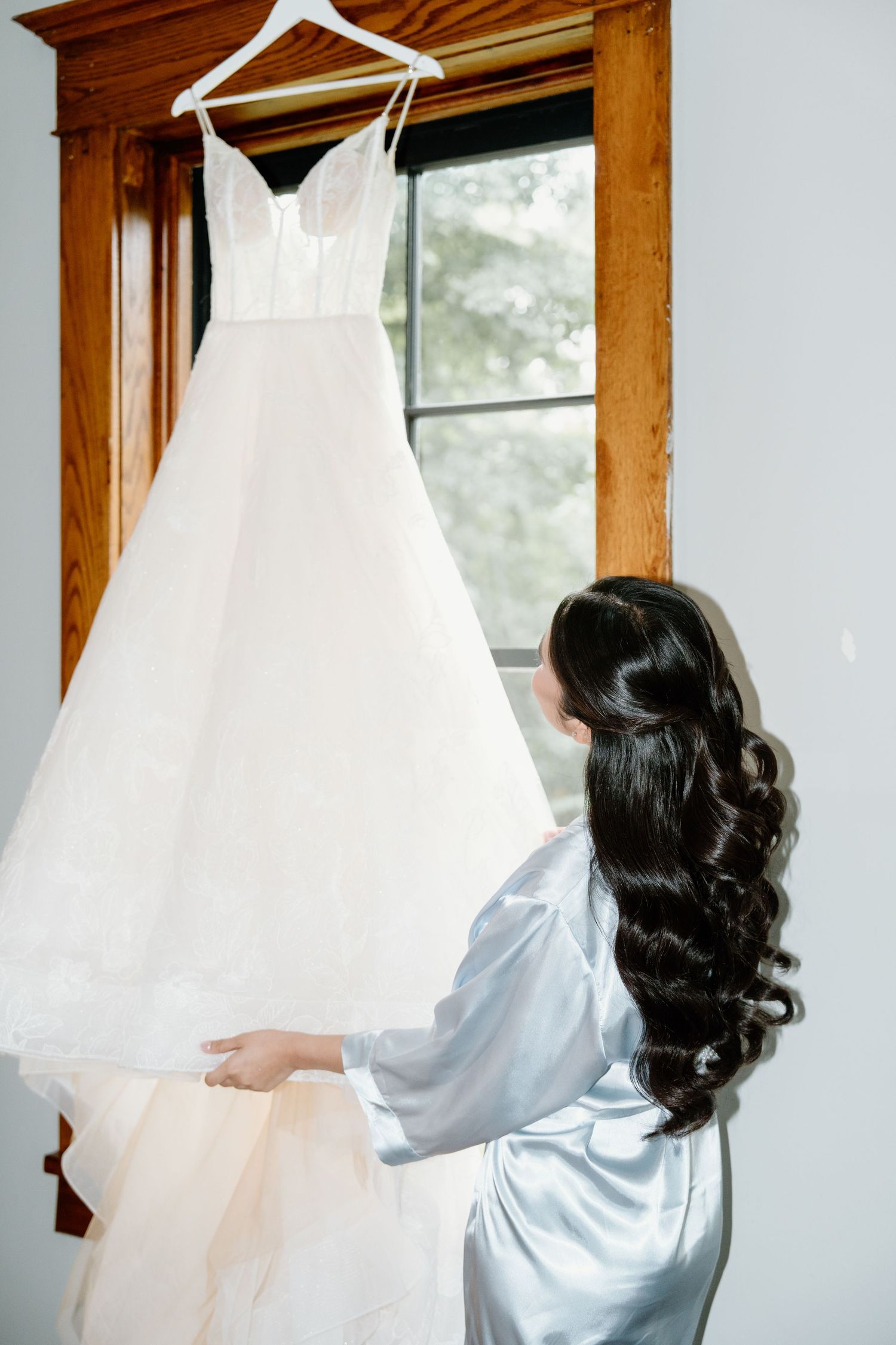 A person in a silk robe looks at white wedding dresses hanging by a window in a bridal preparation room.