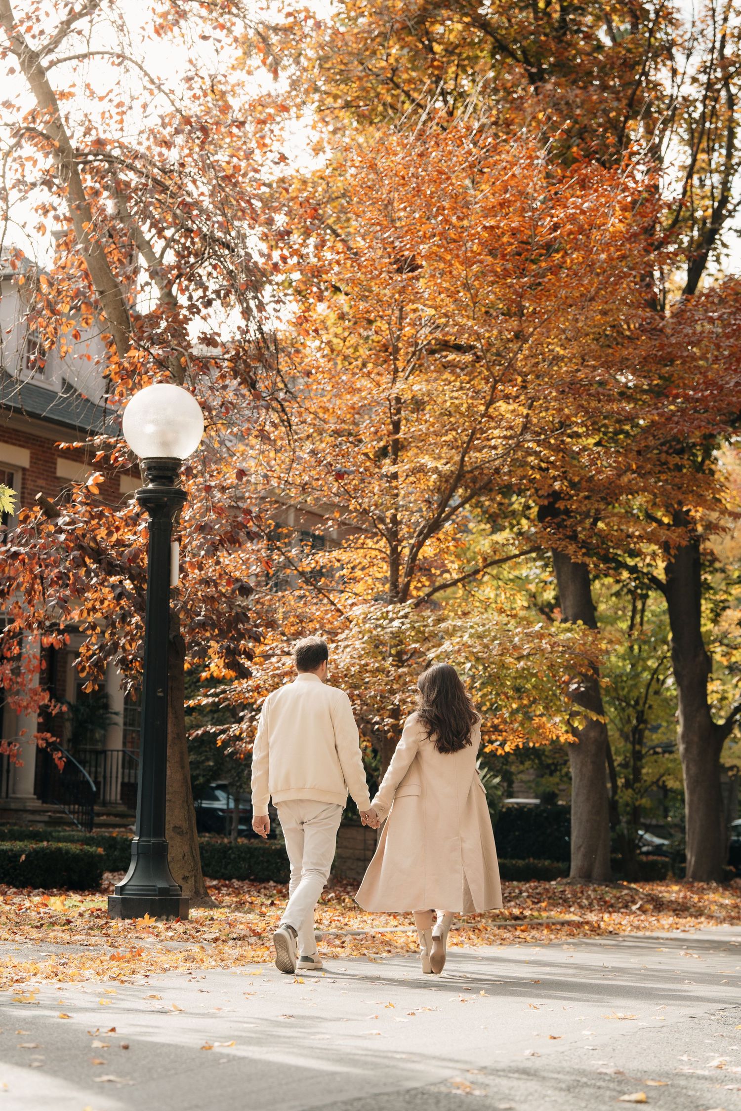 Two people walk along a tree-lined path with autumn foliage and street lamps on a fall day.