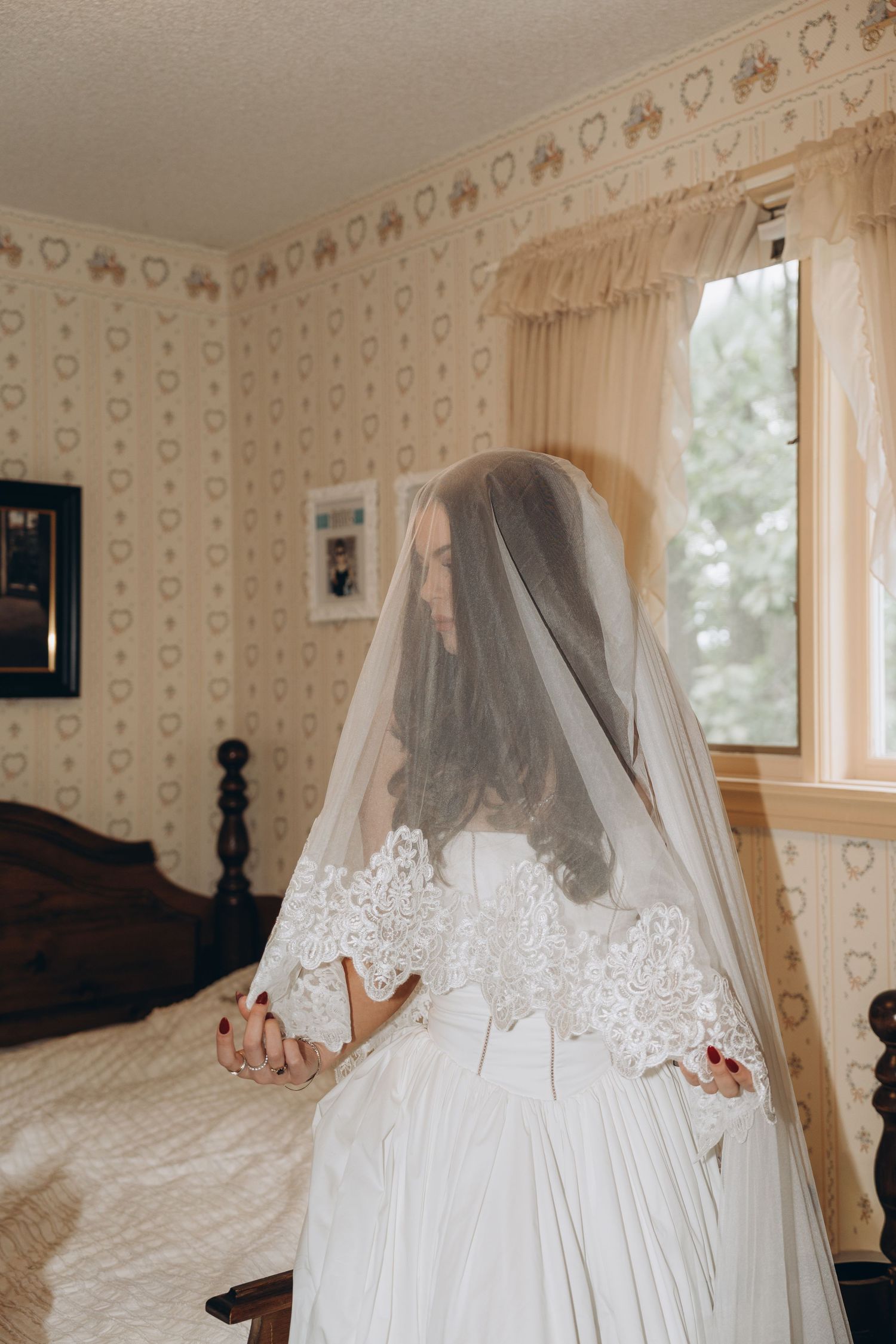 A bride in a flowing white dress and veil poses gracefully near a window in a room with vintage floral wallpaper.