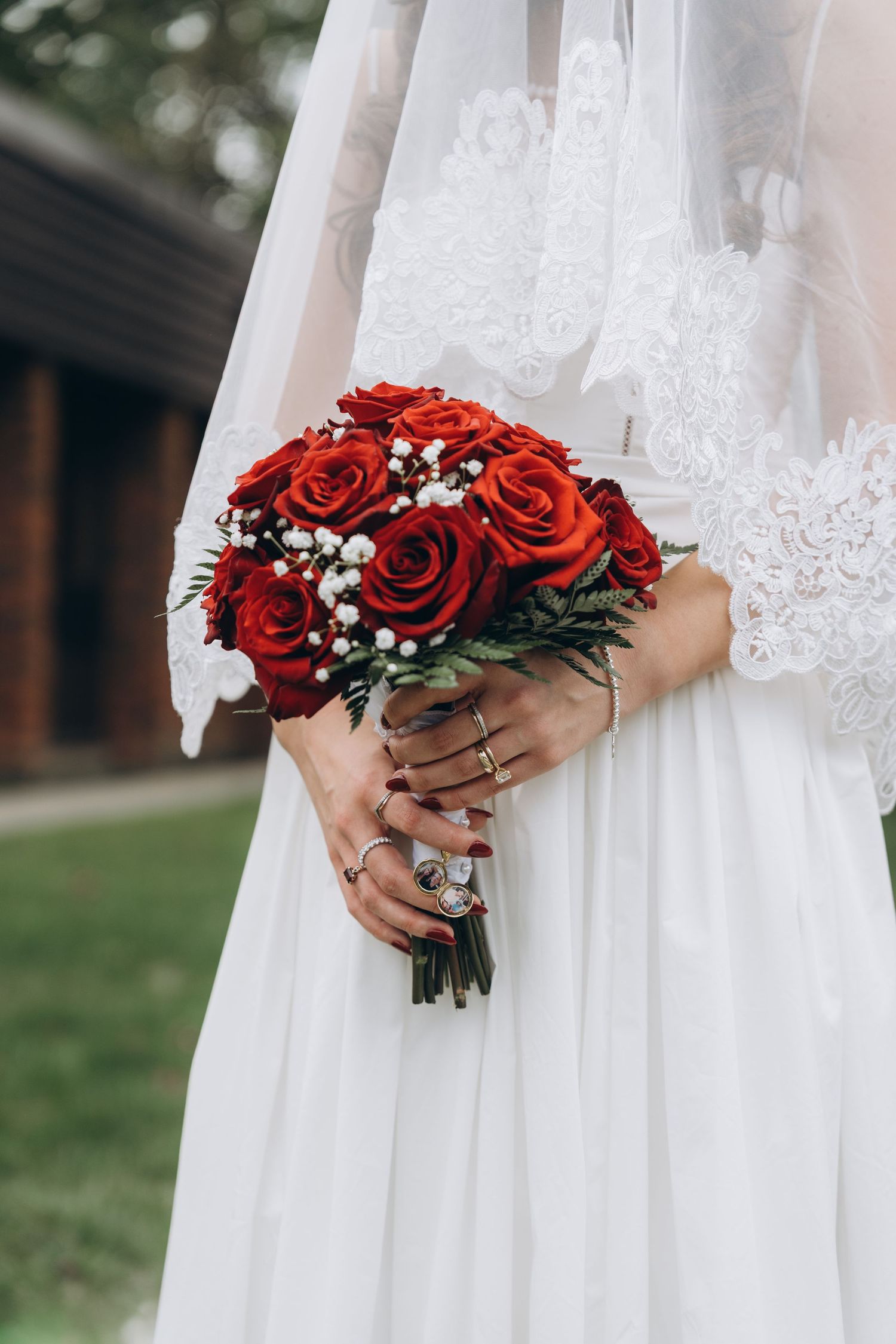 A bride in white lace dress holding a vibrant red rose bouquet with baby's breath against a soft outdoor background.