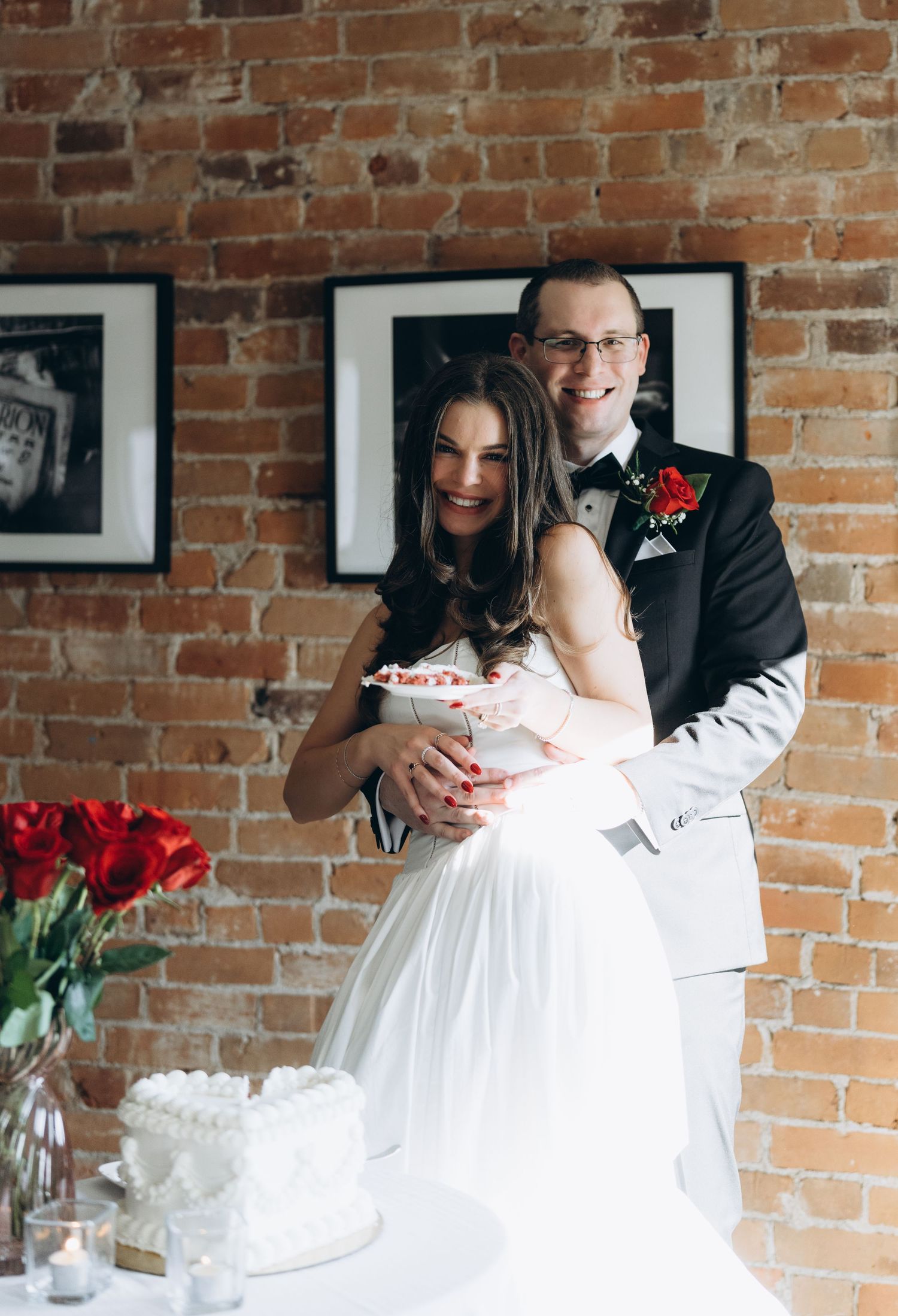 Black and white artistic collage of a wedding couple embracing against exposed brick wall with framed photos and red roses.