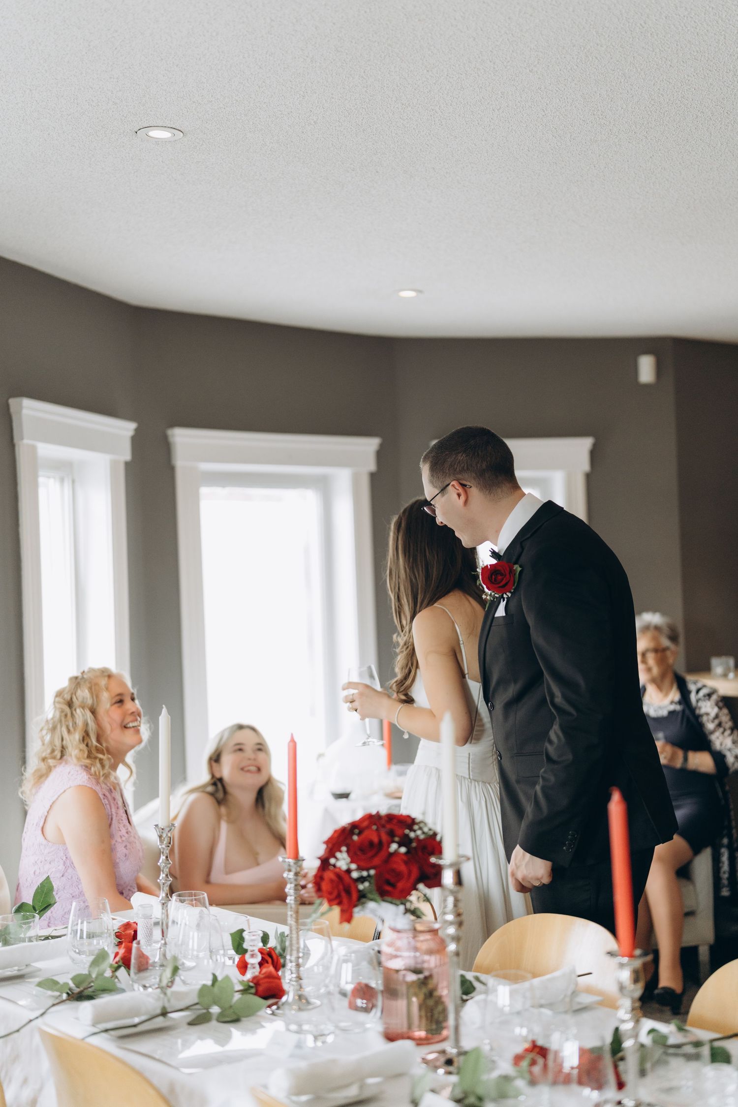 Wedding guests enjoy an elegant dinner reception with red and white decor at long tables by sunlit windows.