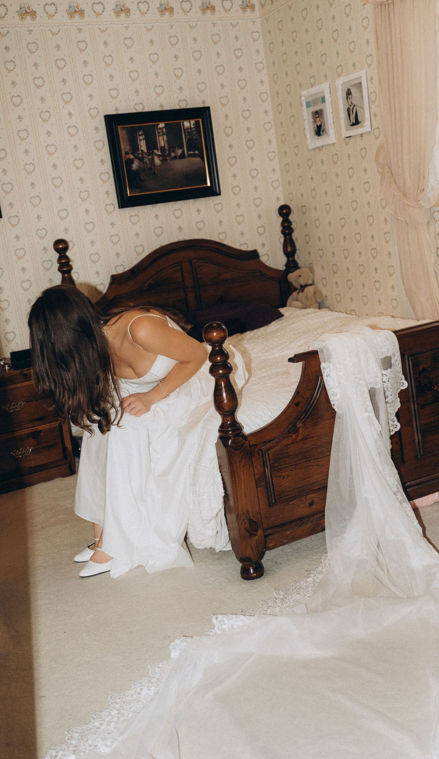 A woman in a white wedding dress sits on the edge of an ornate wooden four-post bed in a vintage styled room.