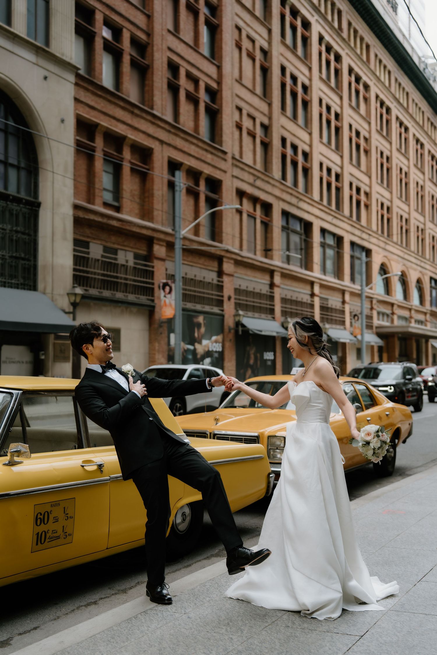 A couple poses playfully in front of a yellow taxi cab on a city street with historic brick buildings in the background.