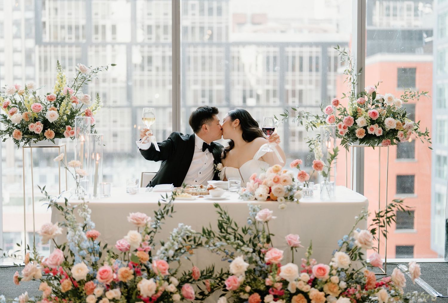 Elegant wedding reception tables decorated with soft pink floral arrangements and tall glass candelabras in a bright venue.