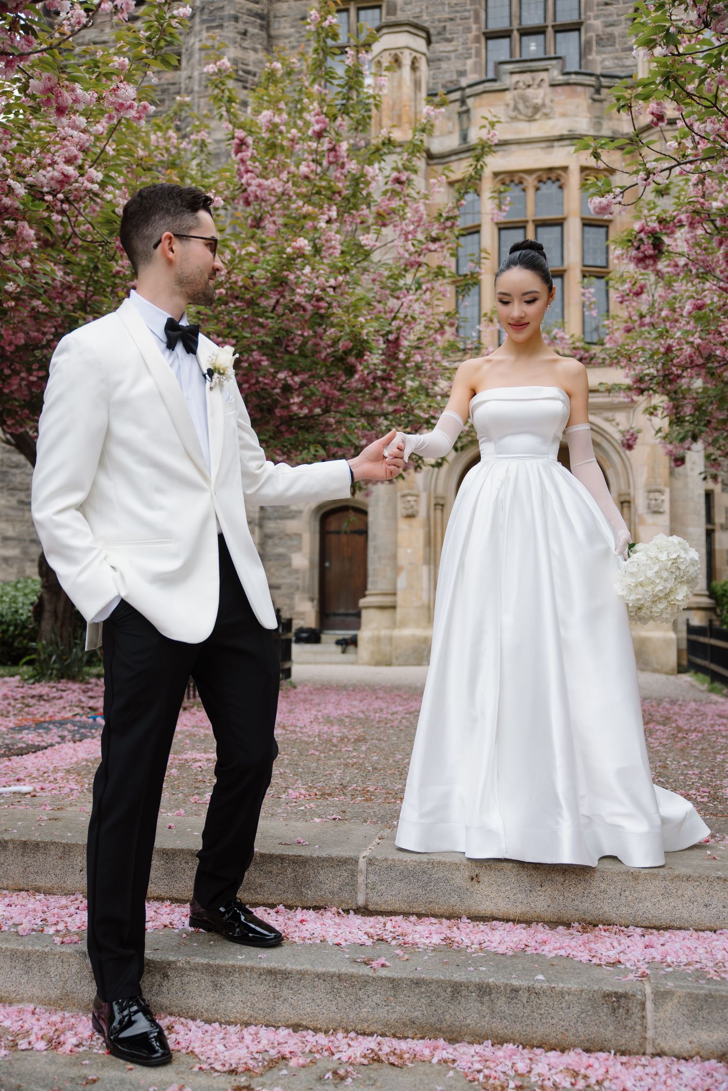 A bride and groom pose on stone steps surrounded by blooming cherry blossoms at a historic building.