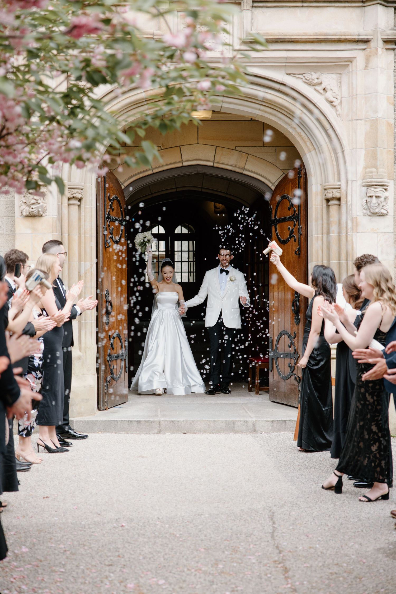 A newlywed couple exits a grand stone archway while guests in black attire shower them with flower petals and confetti.