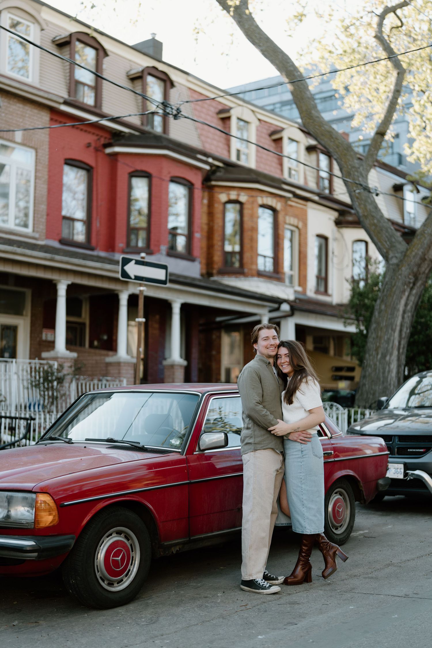 A couple embraces in front of vintage Mercedes on a tree-lined residential street with colorful row houses.