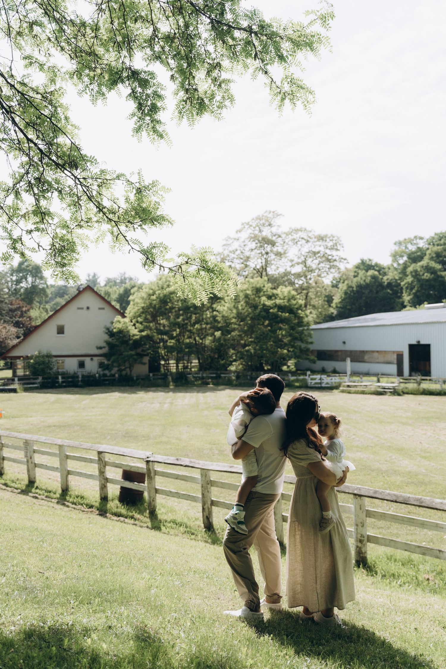 A couple stands together near a wooden fence on a sunny day at a rustic farm property with green trees and buildings.