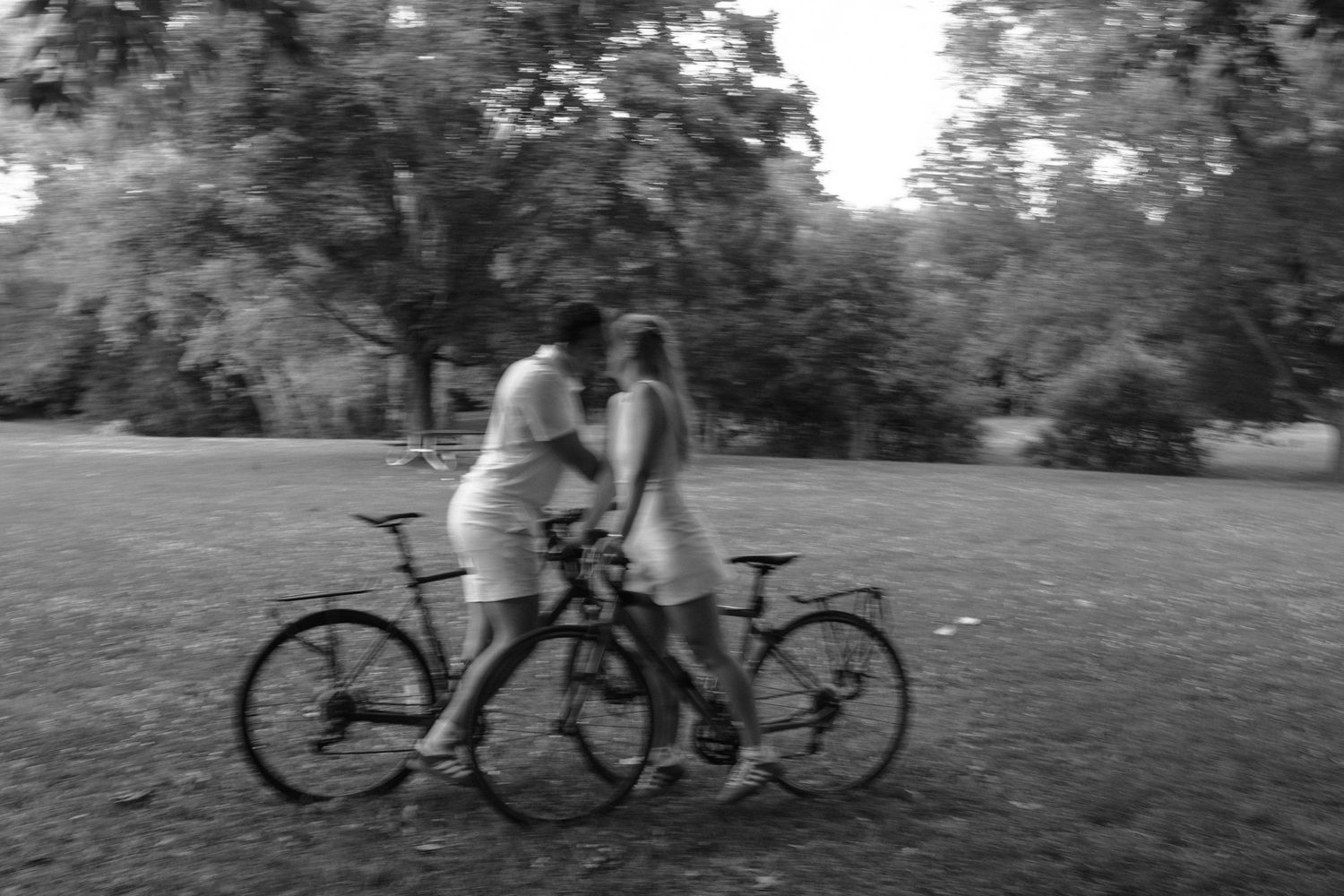 A romantic black and white photo of two cyclists sharing an intimate moment in a park with their bikes.