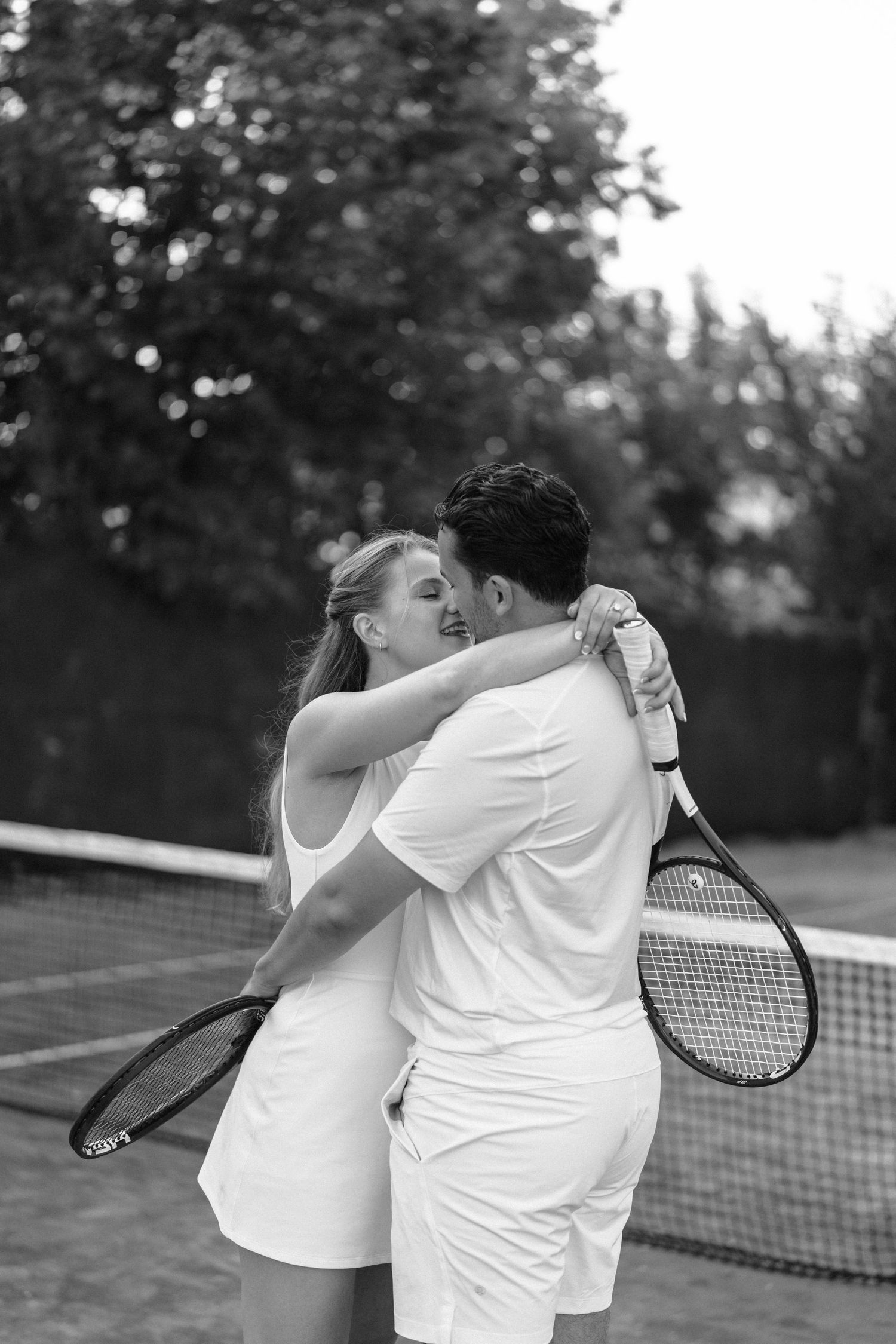 A playful couple in white outfits shares a romantic moment while playing tennis on an outdoor court.