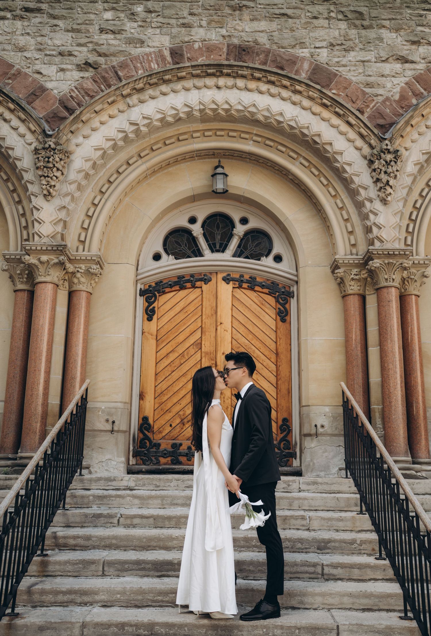A wedding couple stands on stone steps in front of an ornate arched church doorway with romantic architectural details.