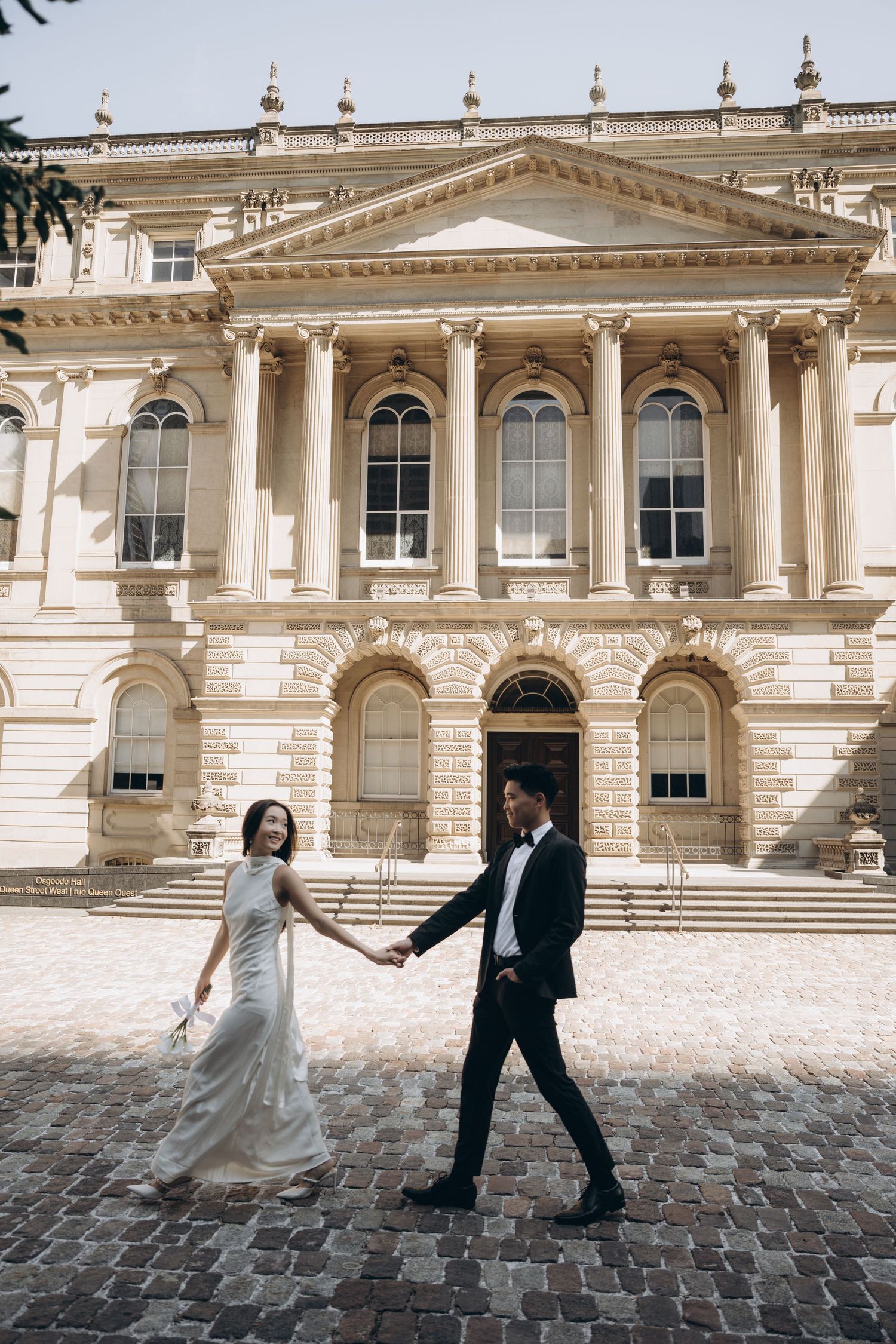 A couple in wedding attire walks on cobblestones in front of a grand neoclassical building with columns and ornate details.