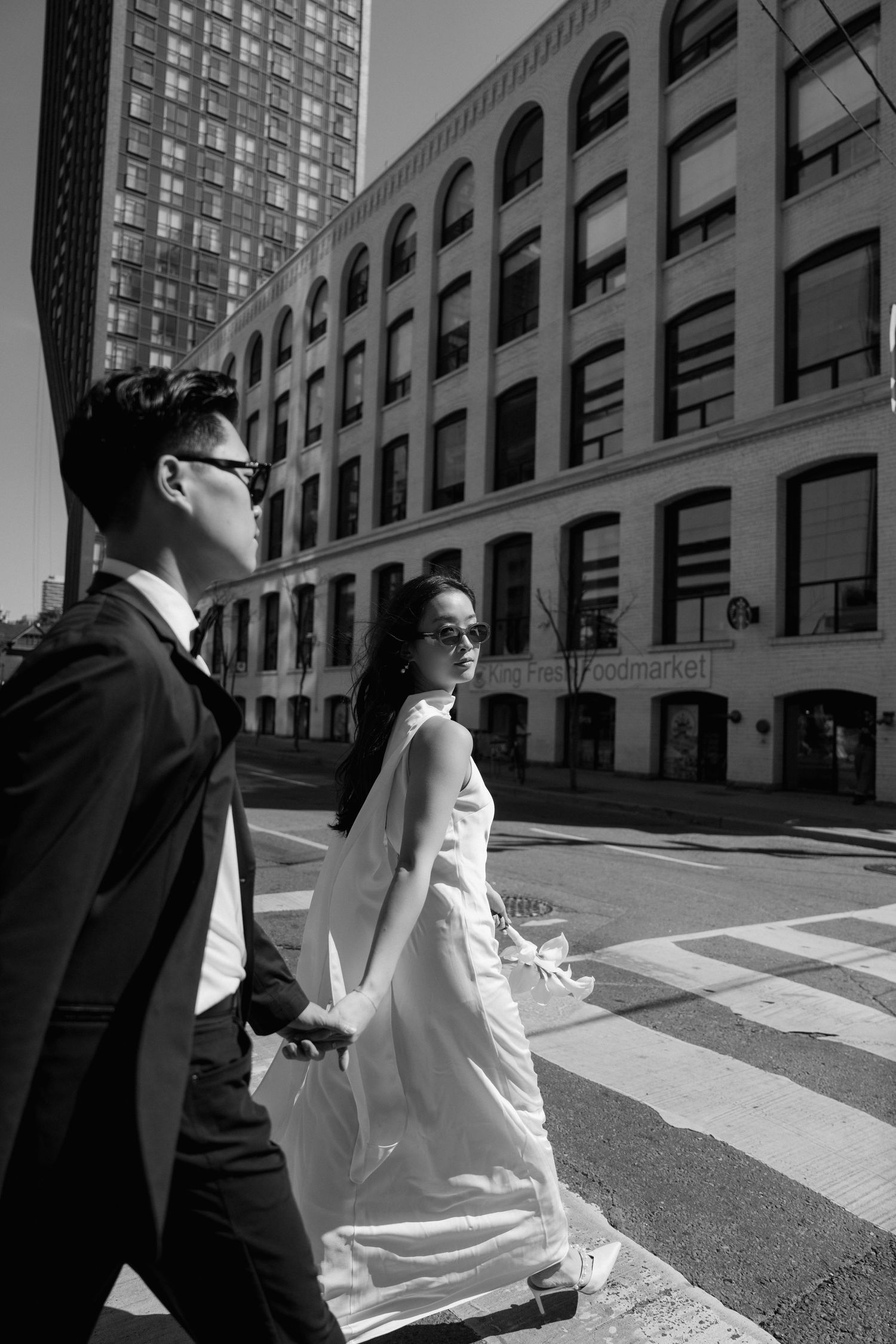 A couple in wedding attire crossing a city street with arched windows and tall buildings in black and white.