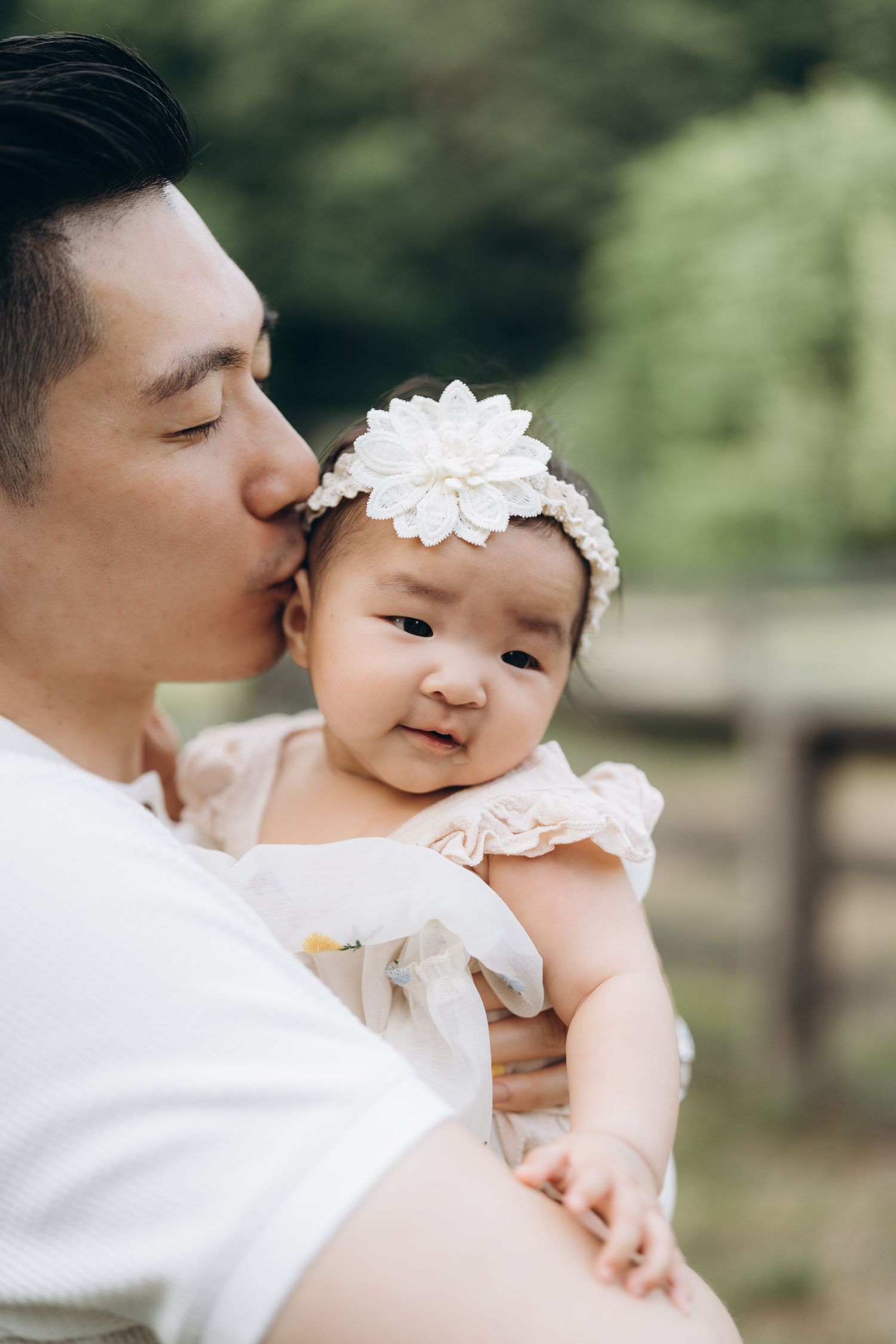 Sweet outdoor family moment as a parent gives loving kisses to a baby wearing a white floral headband.