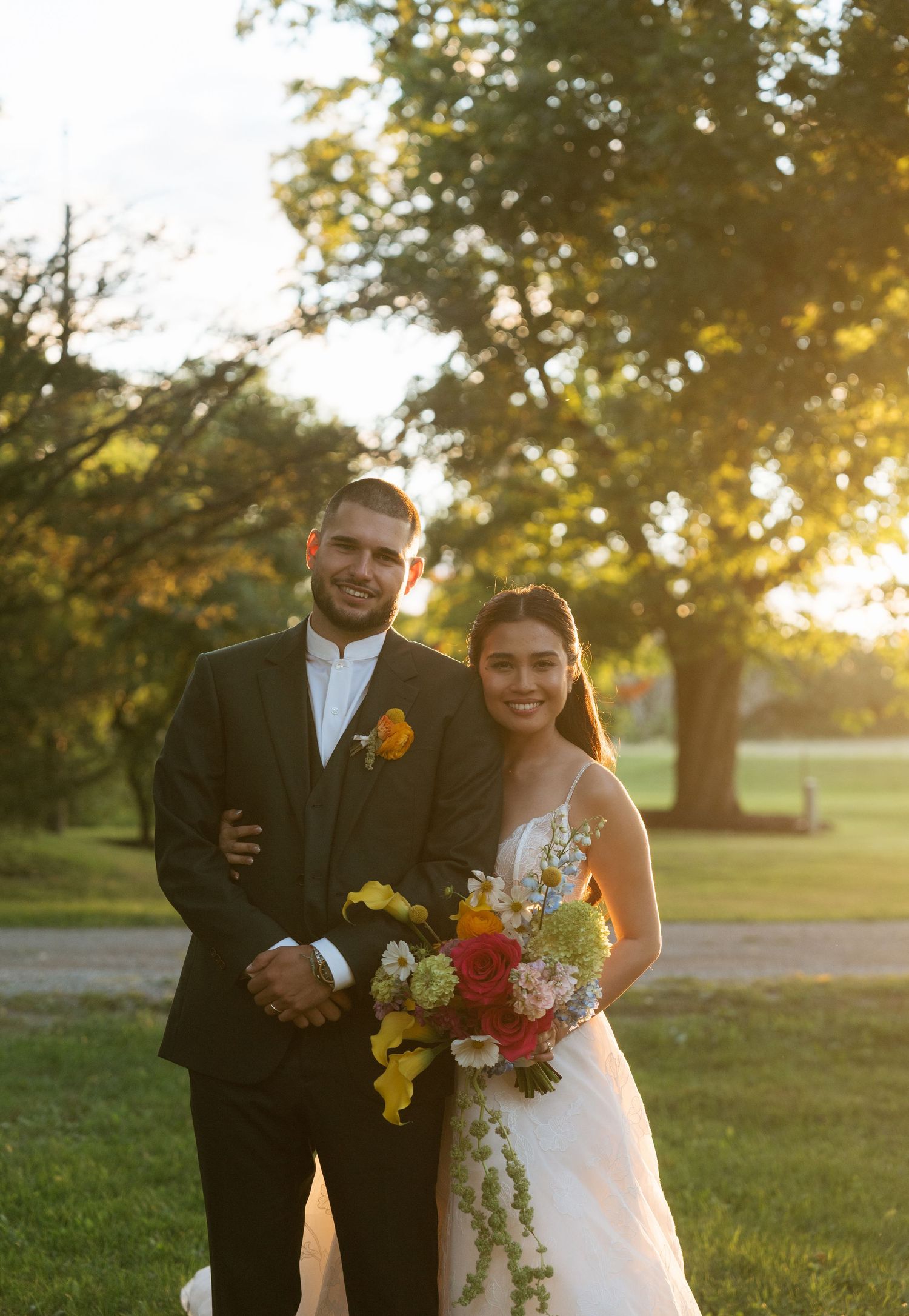 A couple poses together in formal attire during sunset in a lush green park setting.