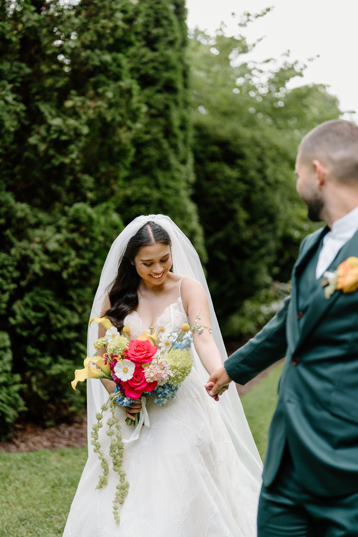 Bride in white dress holding colorful bouquet walks through garden path lined with tall evergreen trees.