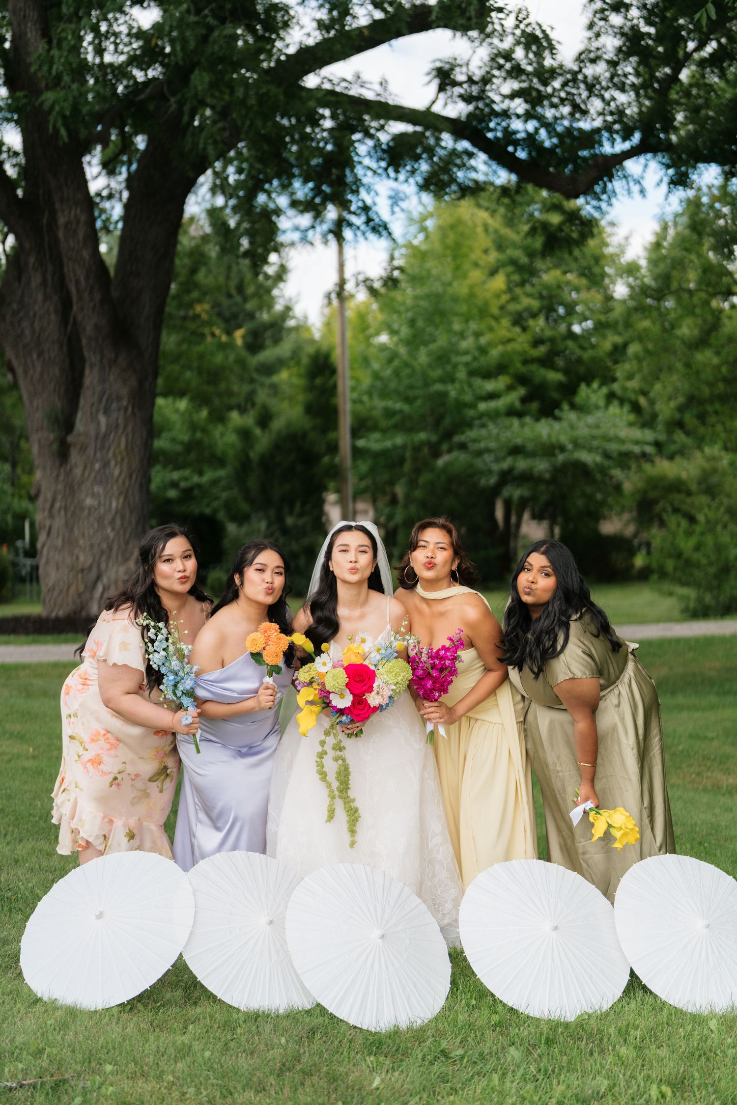 A wedding party poses outdoors with white parasols and colorful bouquets in a garden setting.