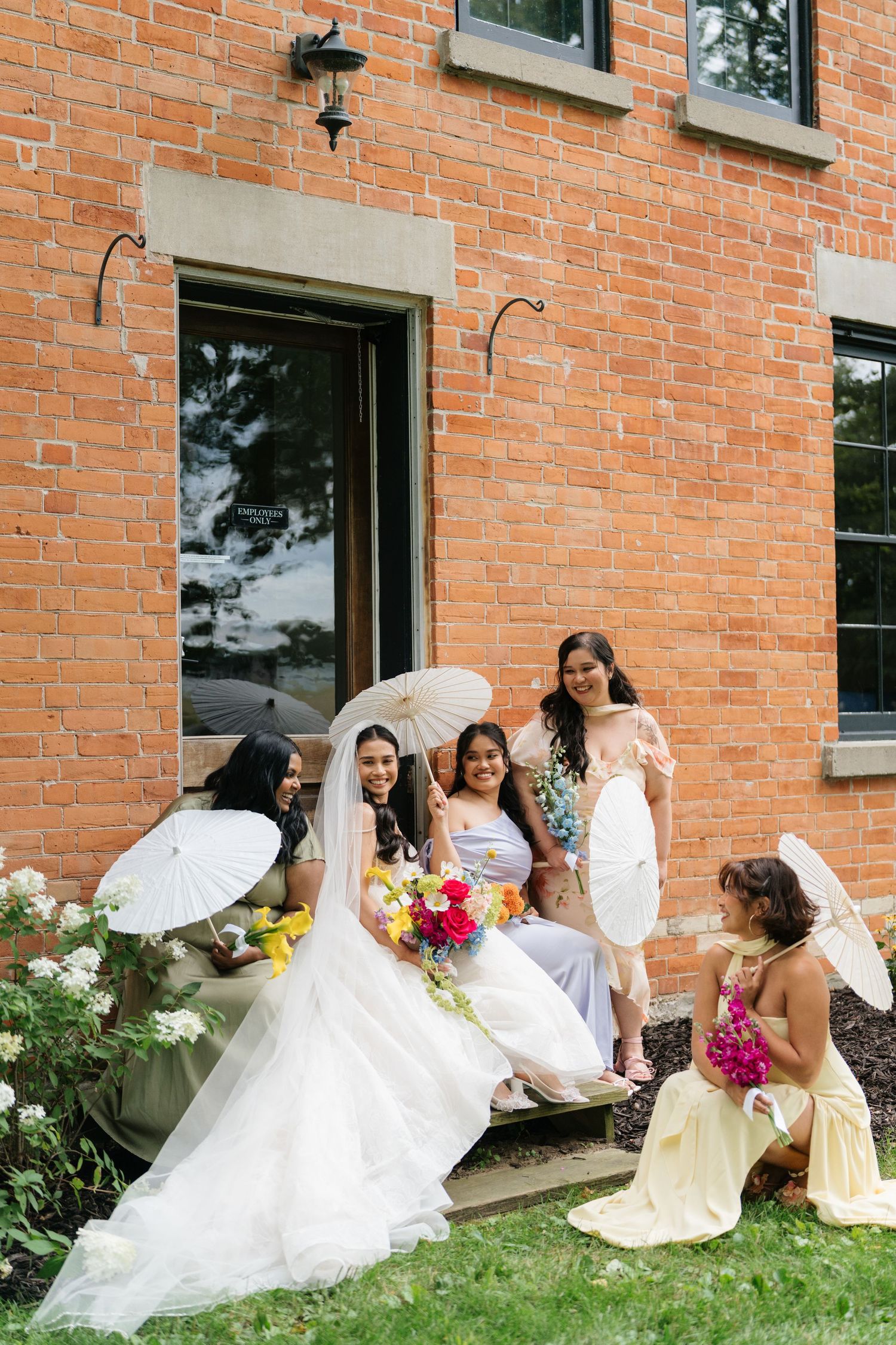 Bridal party members in yellow and white dresses arrange themselves against a red brick wall for photos.