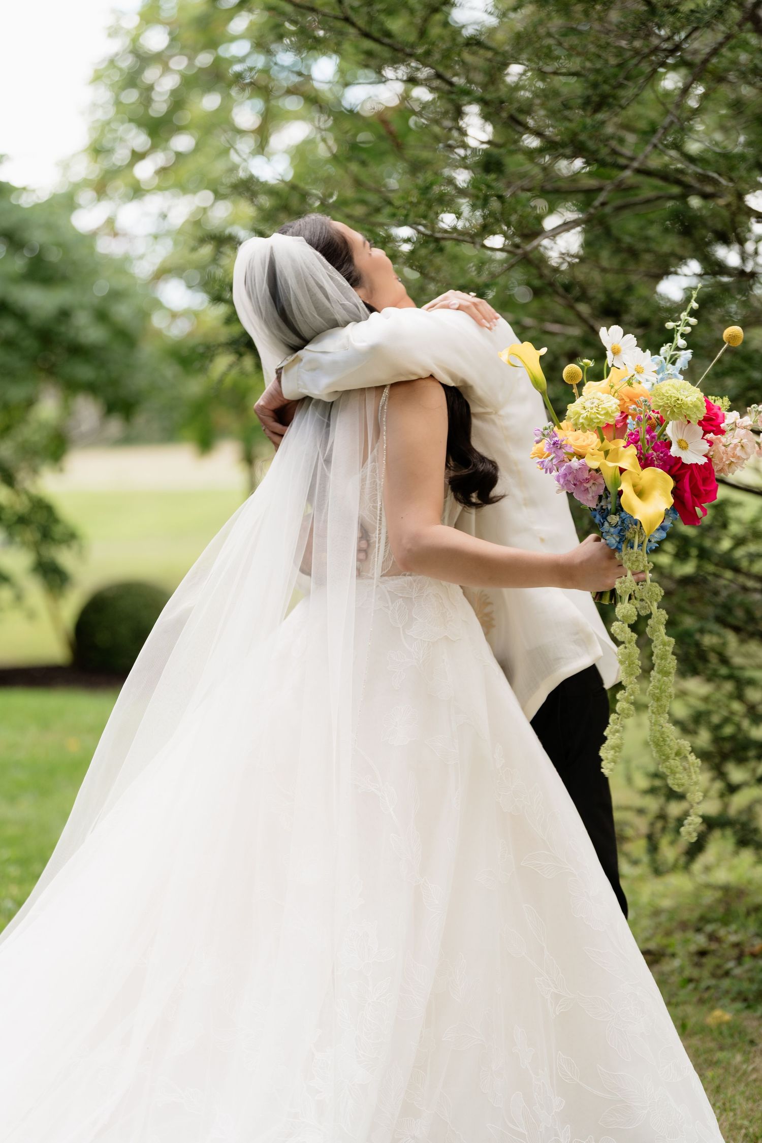 A romantic wedding couple embraces outdoors under flowering trees with a colorful bouquet of spring flowers.