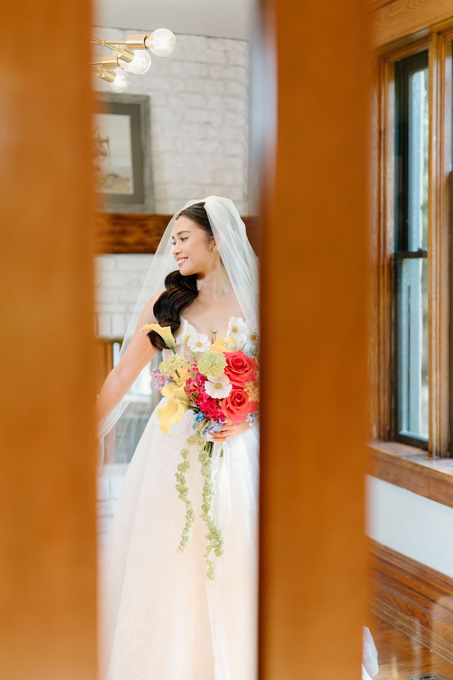 A bride in a white wedding dress and veil holds a vibrant coral and peach floral bouquet against a rustic indoor backdrop.