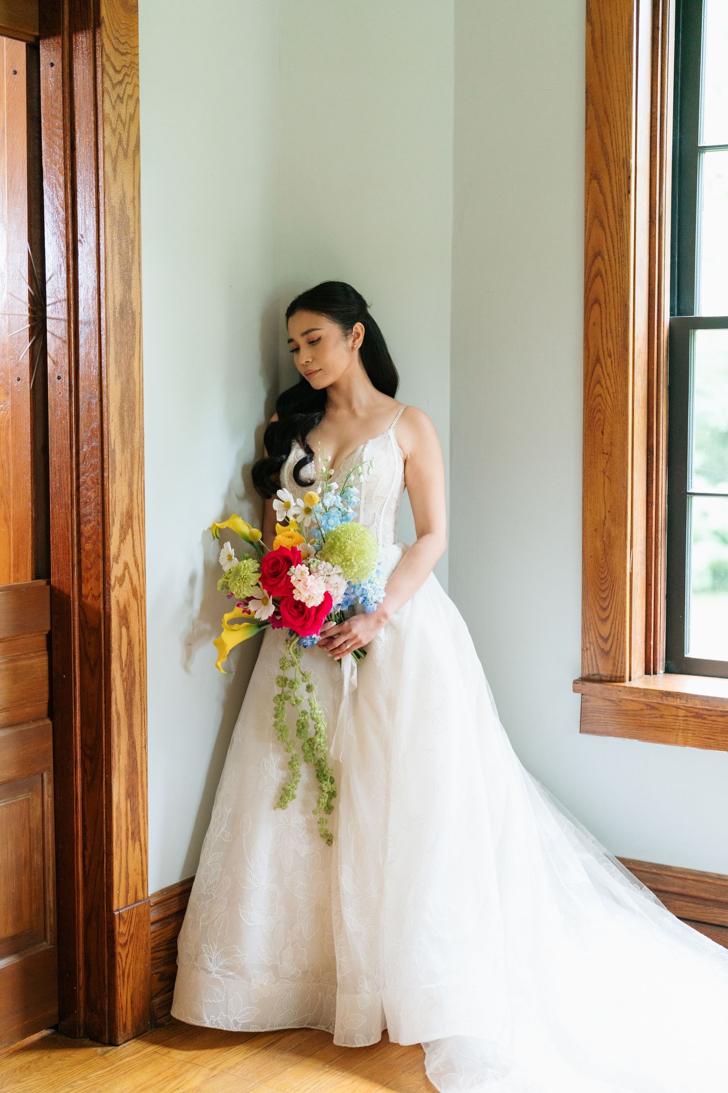 Elegant bride in white ballgown wedding dress holds vibrant bouquet with yellow lilies and pink flowers by wooden window frame.